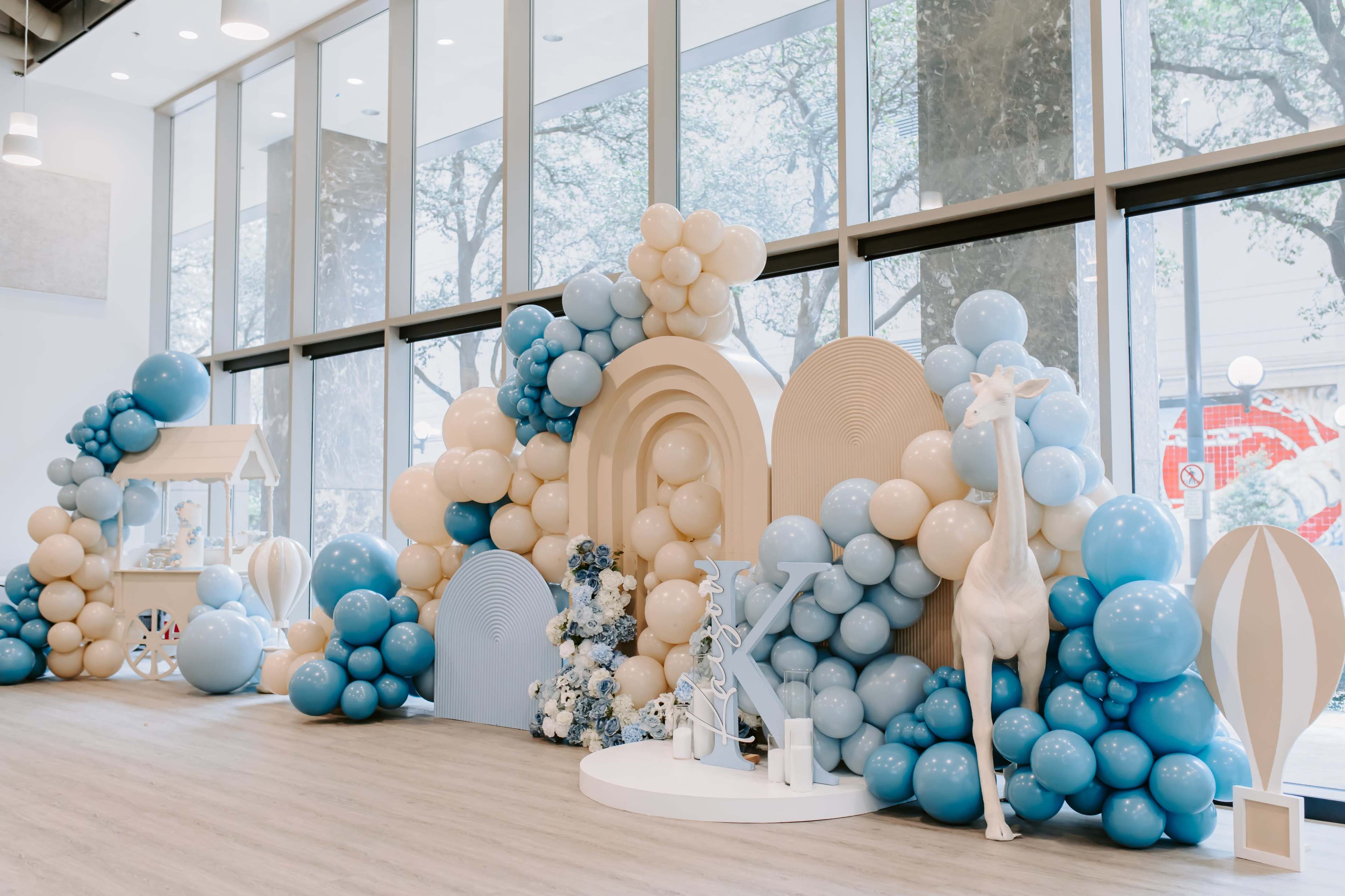 The scene features a decorative backdrop made of blue and beige balloons arranged around geometric shapes in a brightly lit indoor space with large windows.