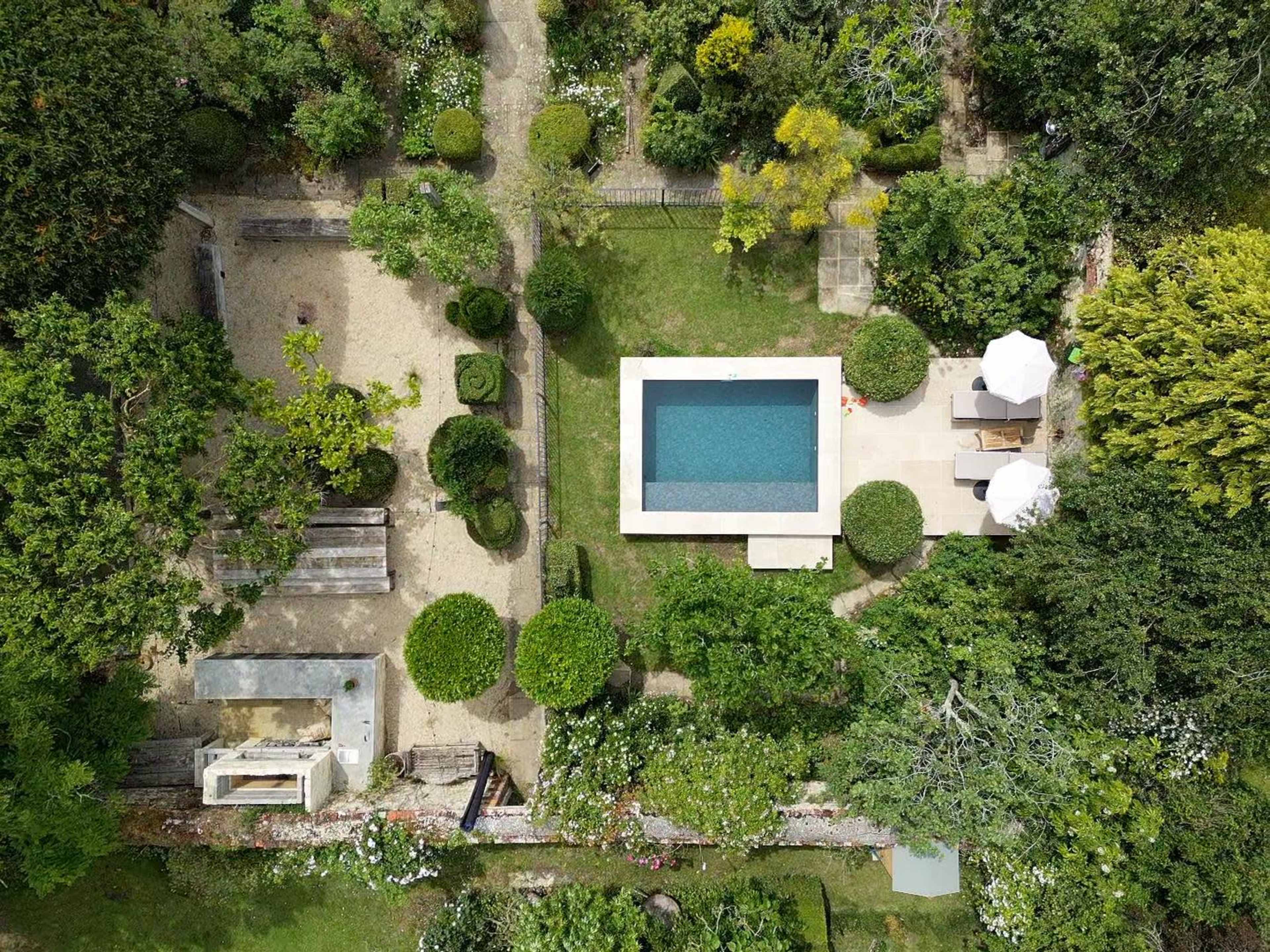 The image shows an aerial view of a landscaped garden featuring a swimming pool, manicured hedges, and a seating area with umbrellas.
