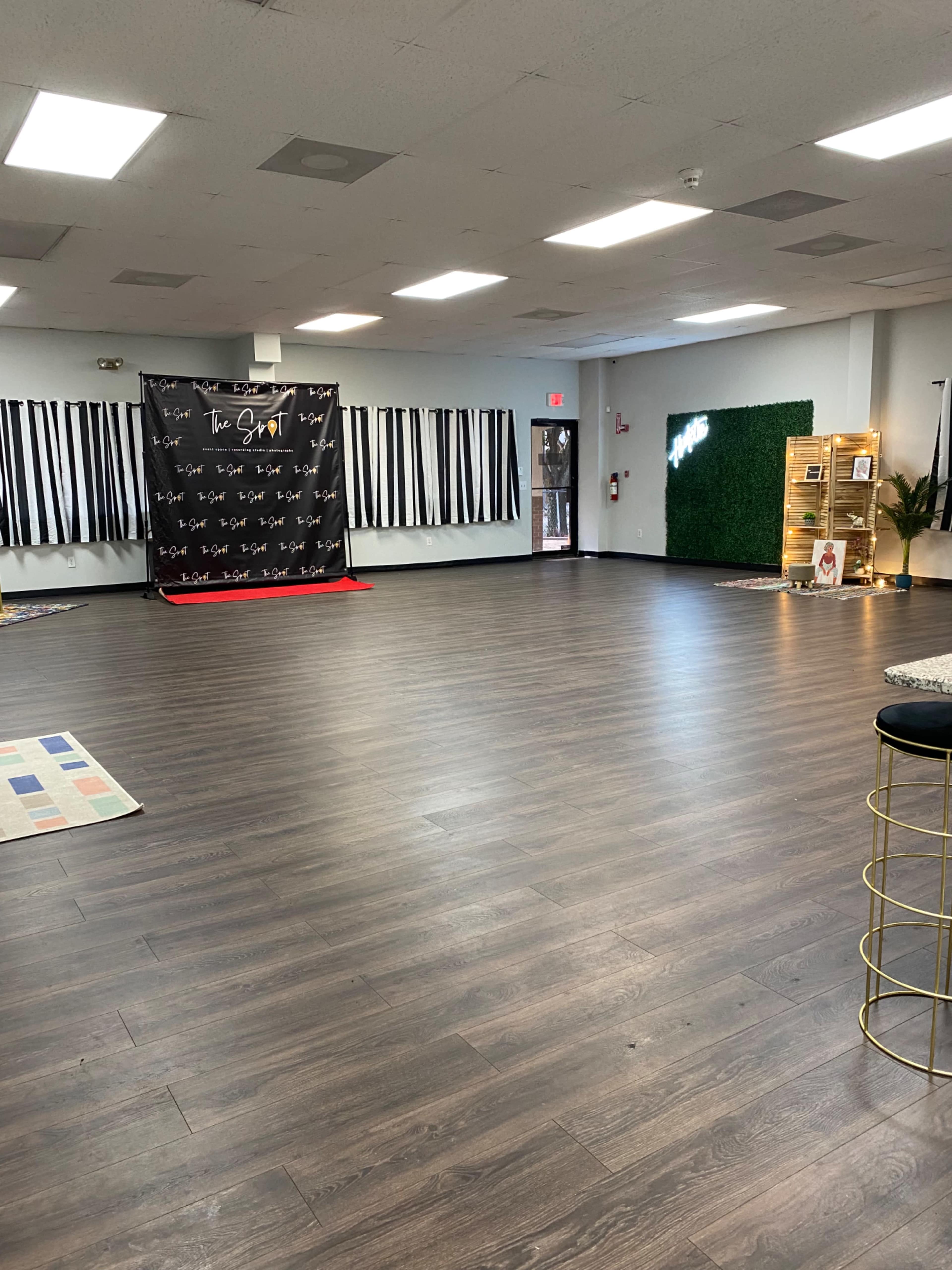 A spacious room with wooden flooring, decorated with black and white striped curtains, a backdrop for an event, and a small area with greenery and furniture.