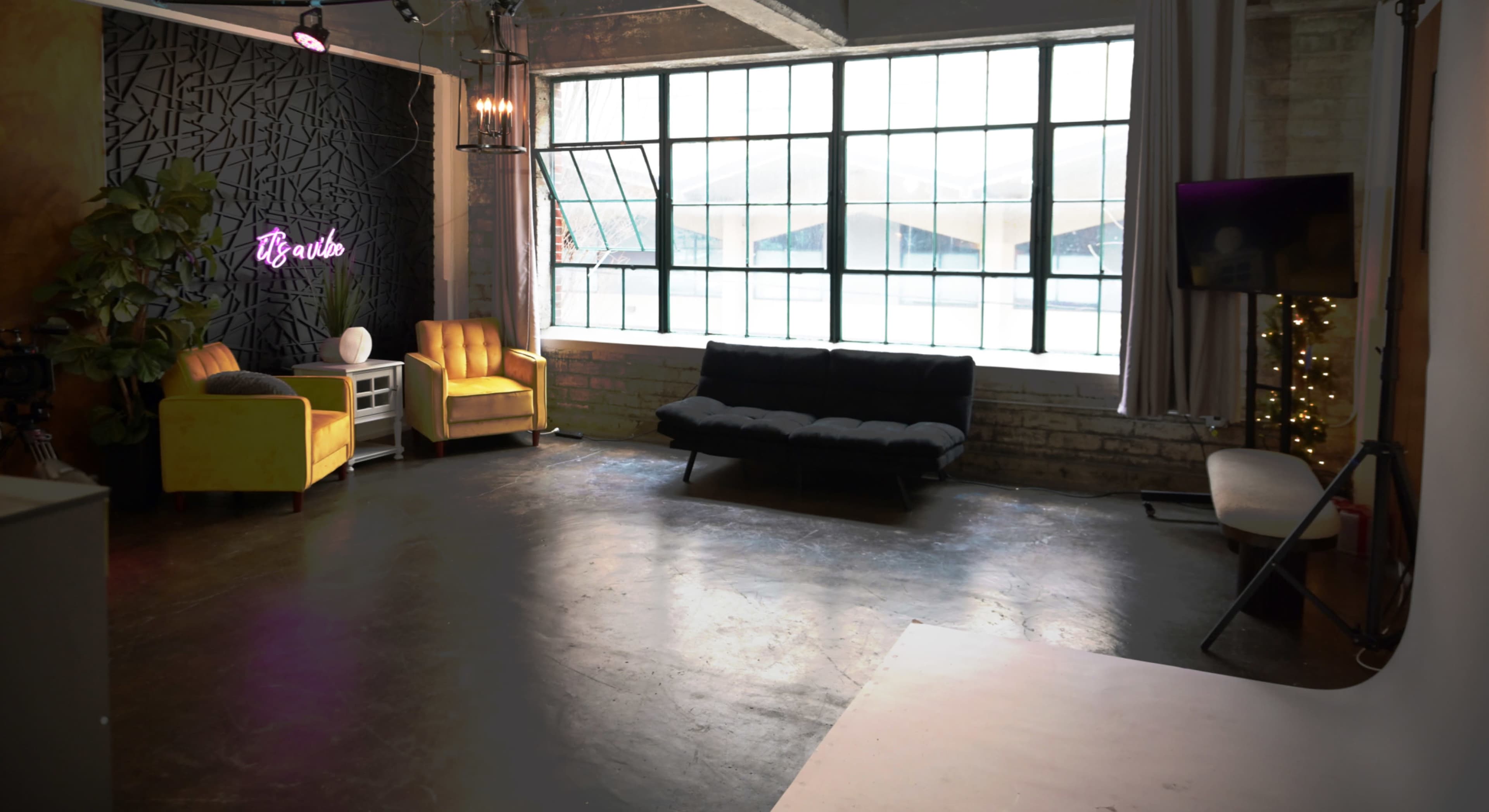 The image shows a spacious interior featuring two yellow chairs, a black couch, large windows, and a wall with a decorative design and neon lights.