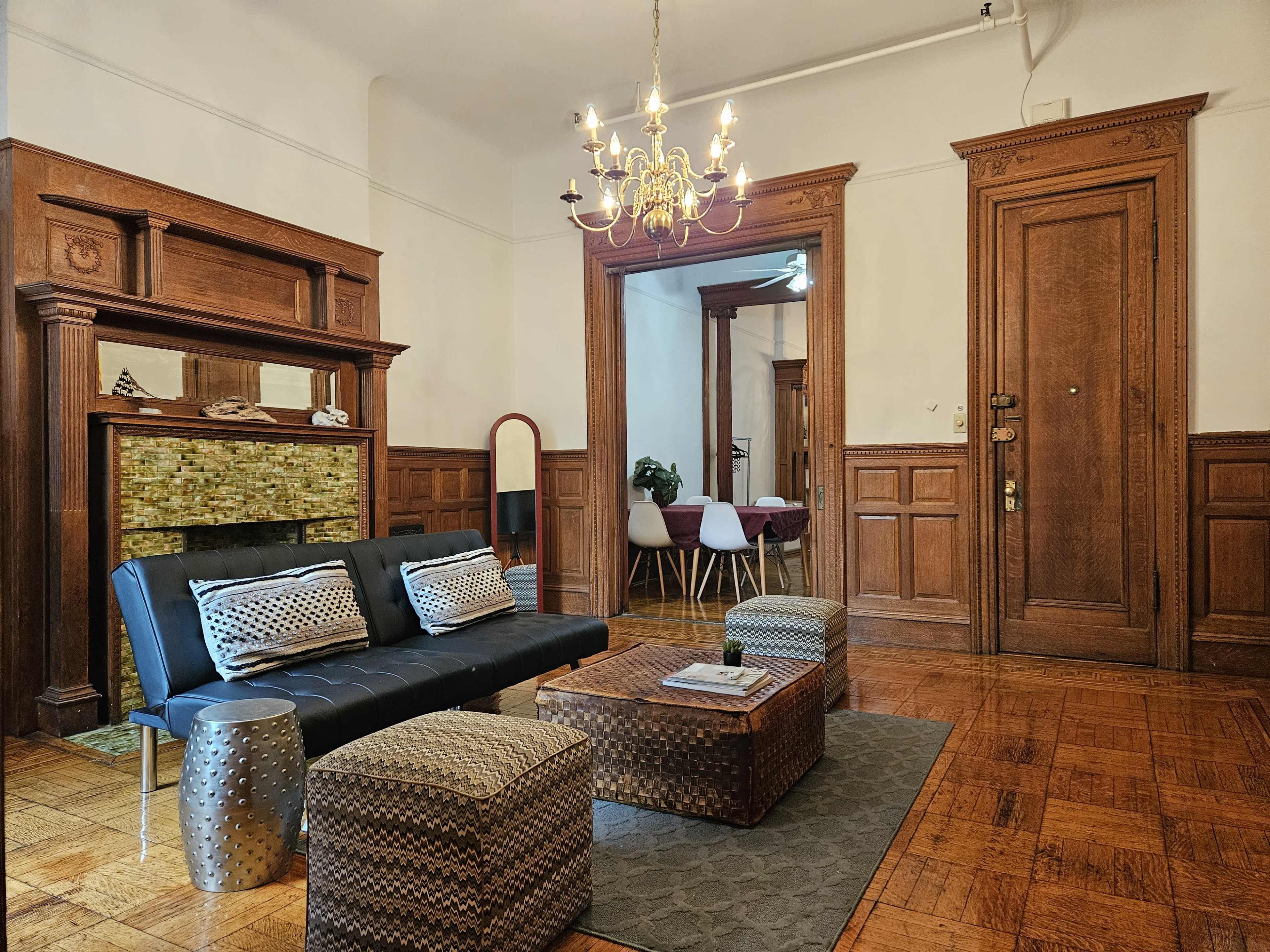 A living room features a black sofa and a wooden coffee table, surrounded by decorative stools and a chandelier, with paneled walls and a stylish fireplace.