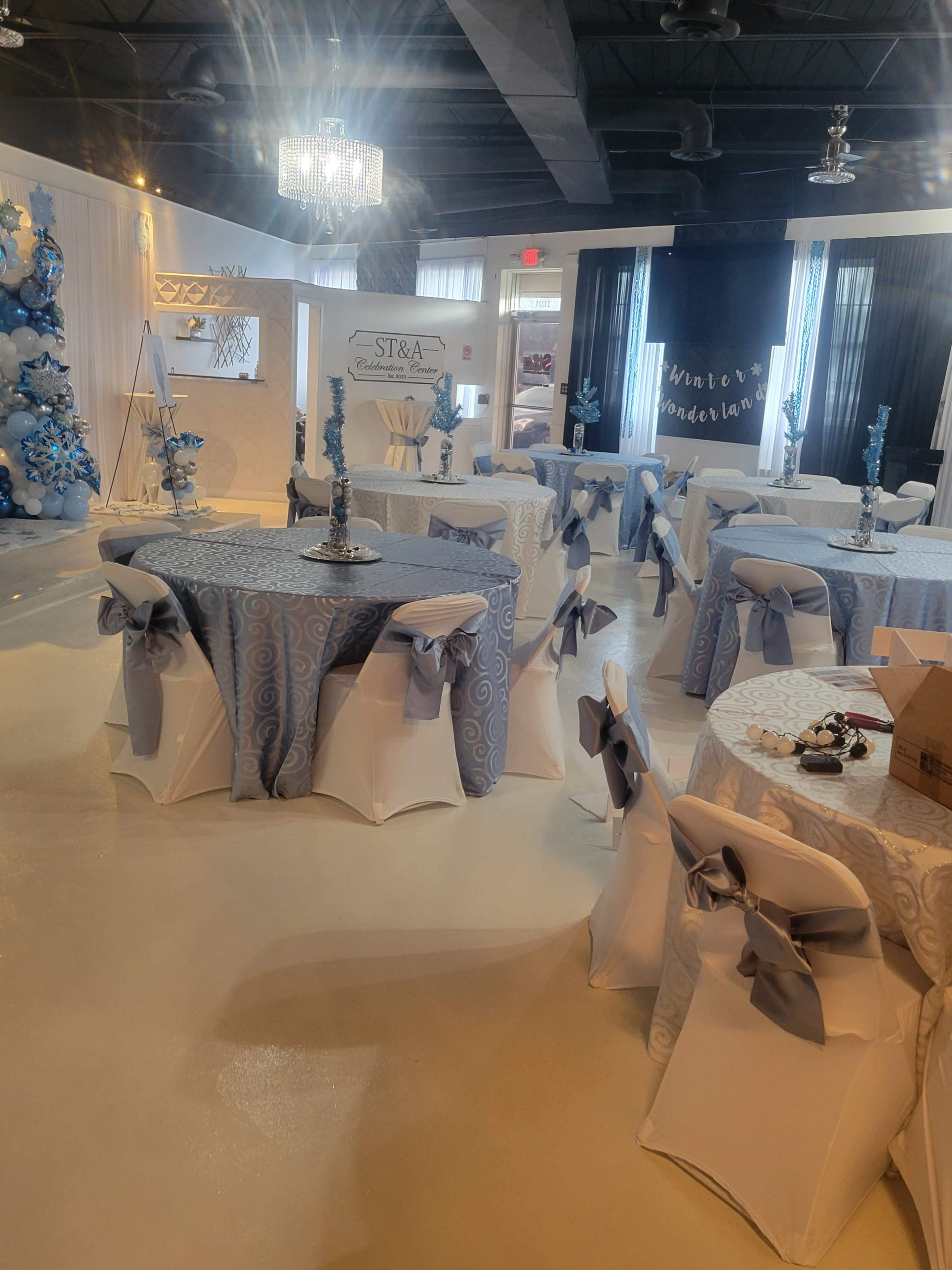 The image shows a decorated event space with tables covered in light blue and white tablecloths, each adorned with a silver candelabra, and elegant chairs draped with matching bows.