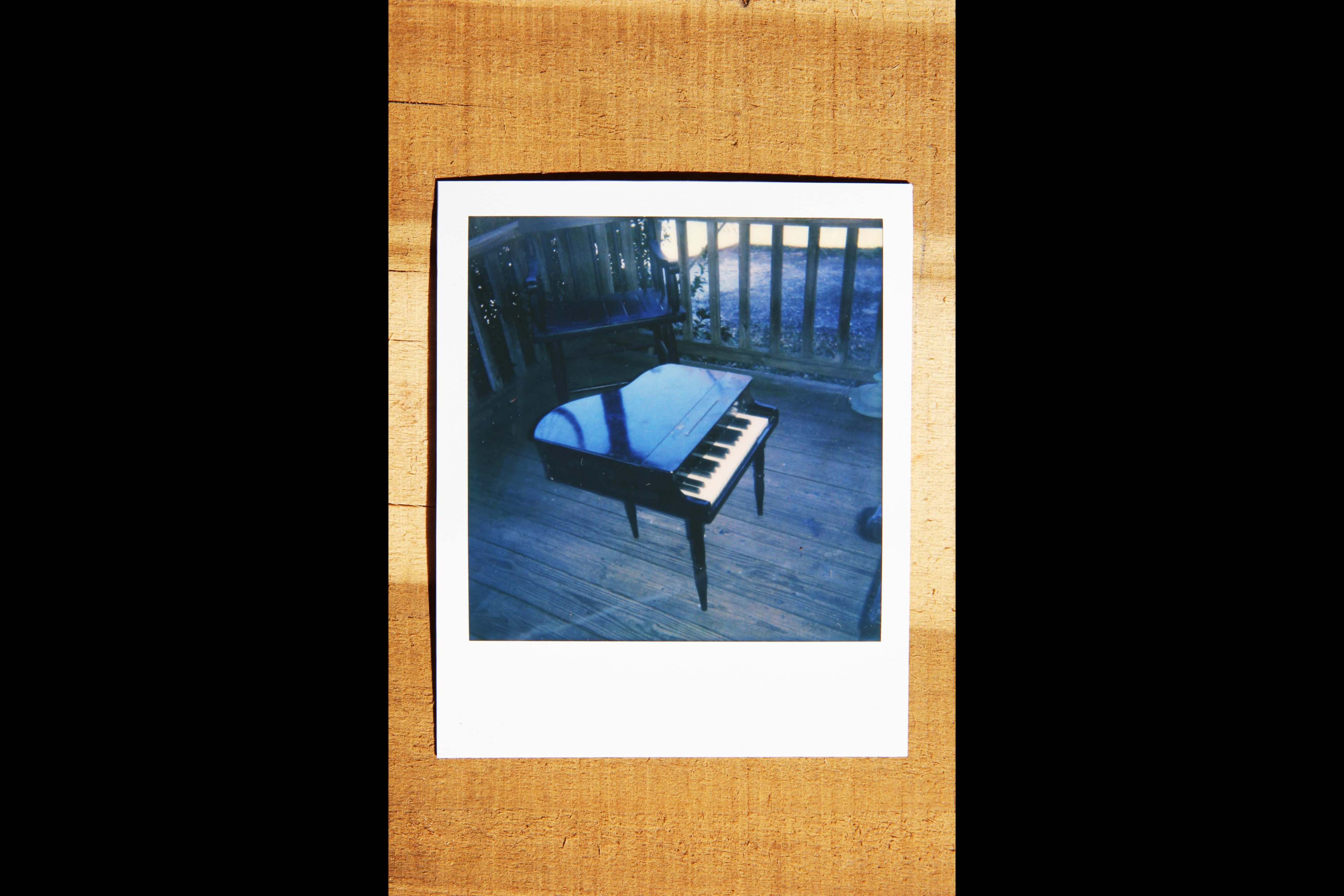 A small piano sits on a wooden porch, casting reflections on its surface.