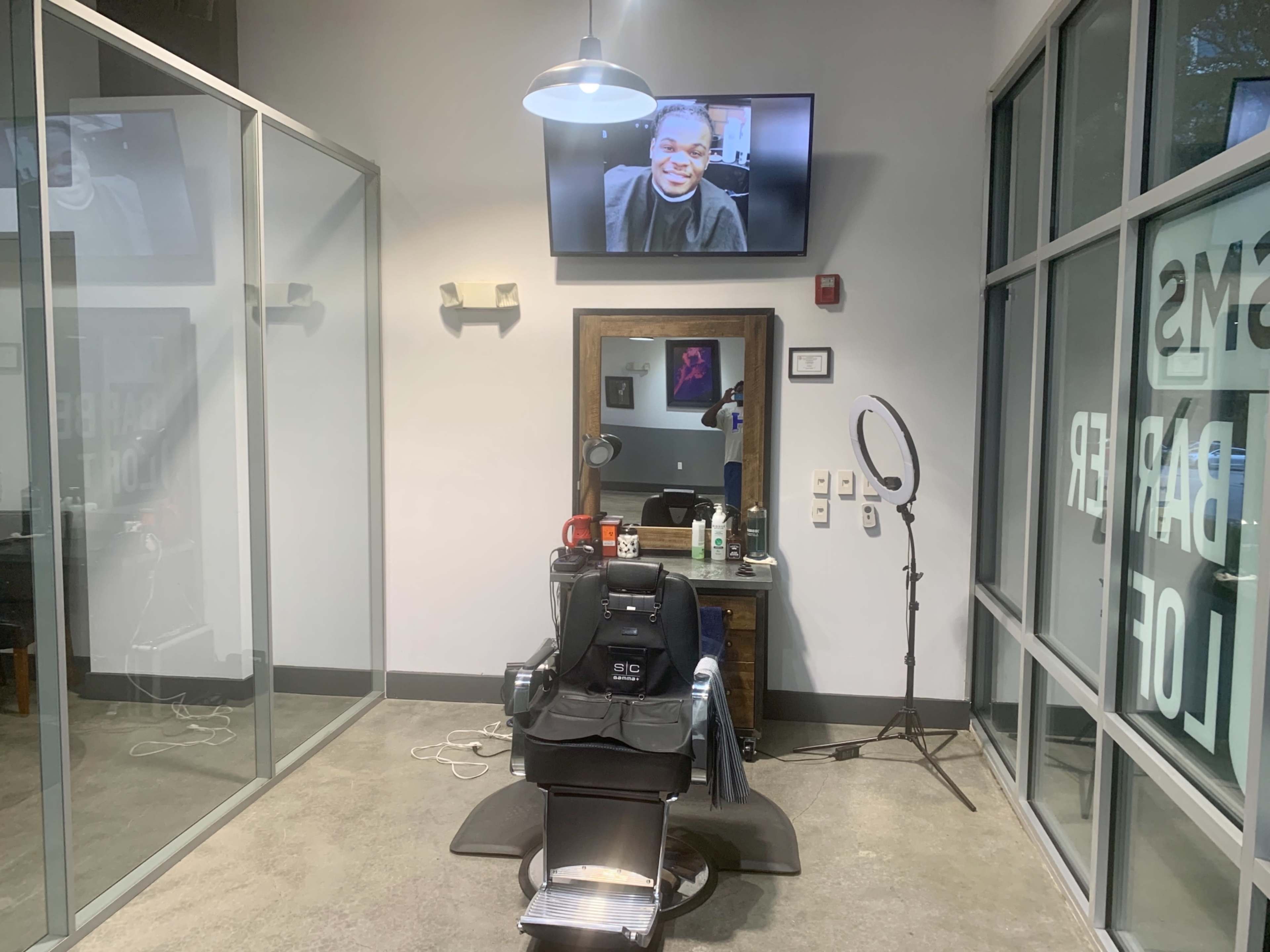 The image shows a modern barbershop interior featuring a black barber chair, a mirror, and a television displaying a person, with large windows and bright lighting.