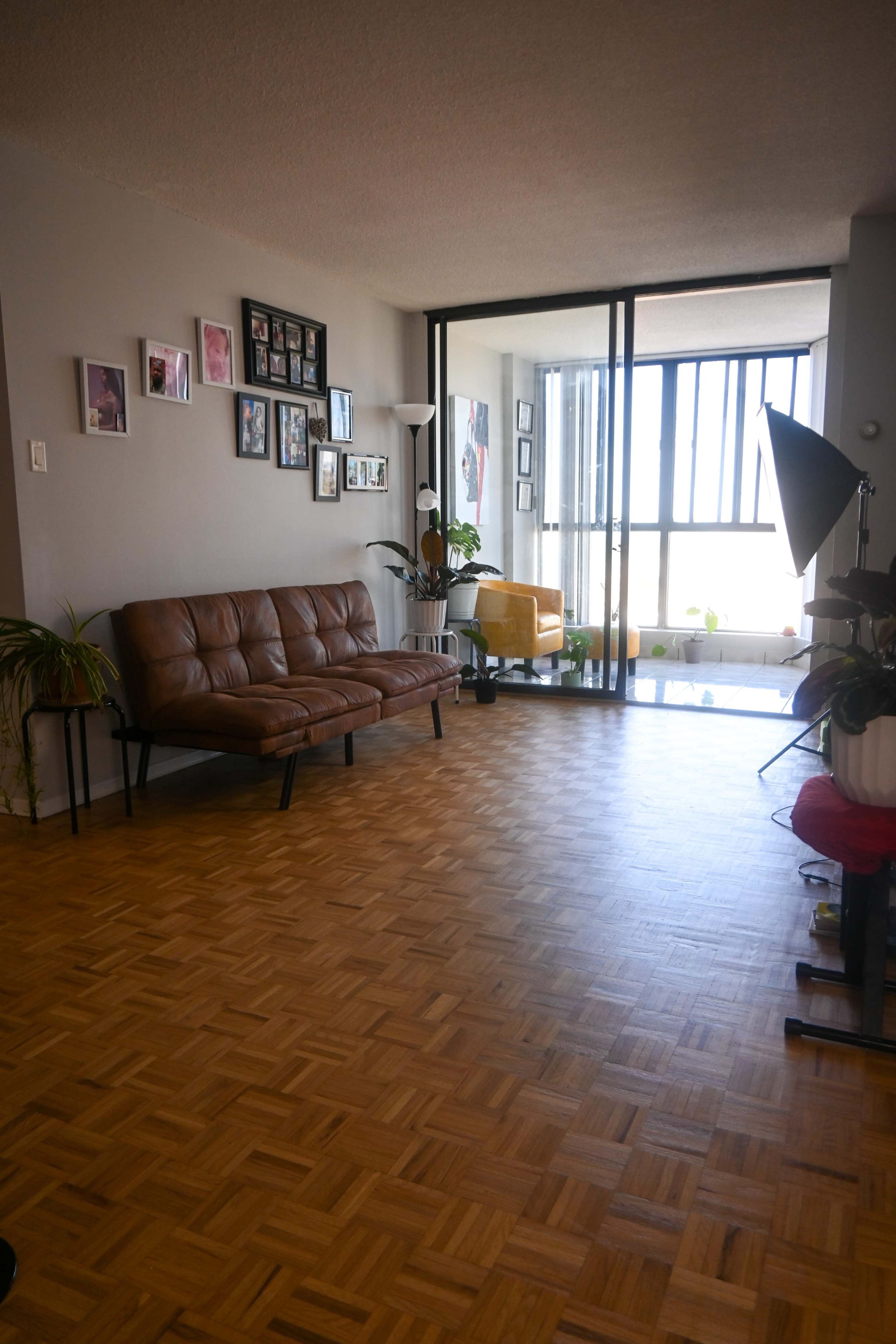 The image shows a living room featuring a brown mid-century sofa, a wall with framed photos, and large windows allowing natural light to enter.
