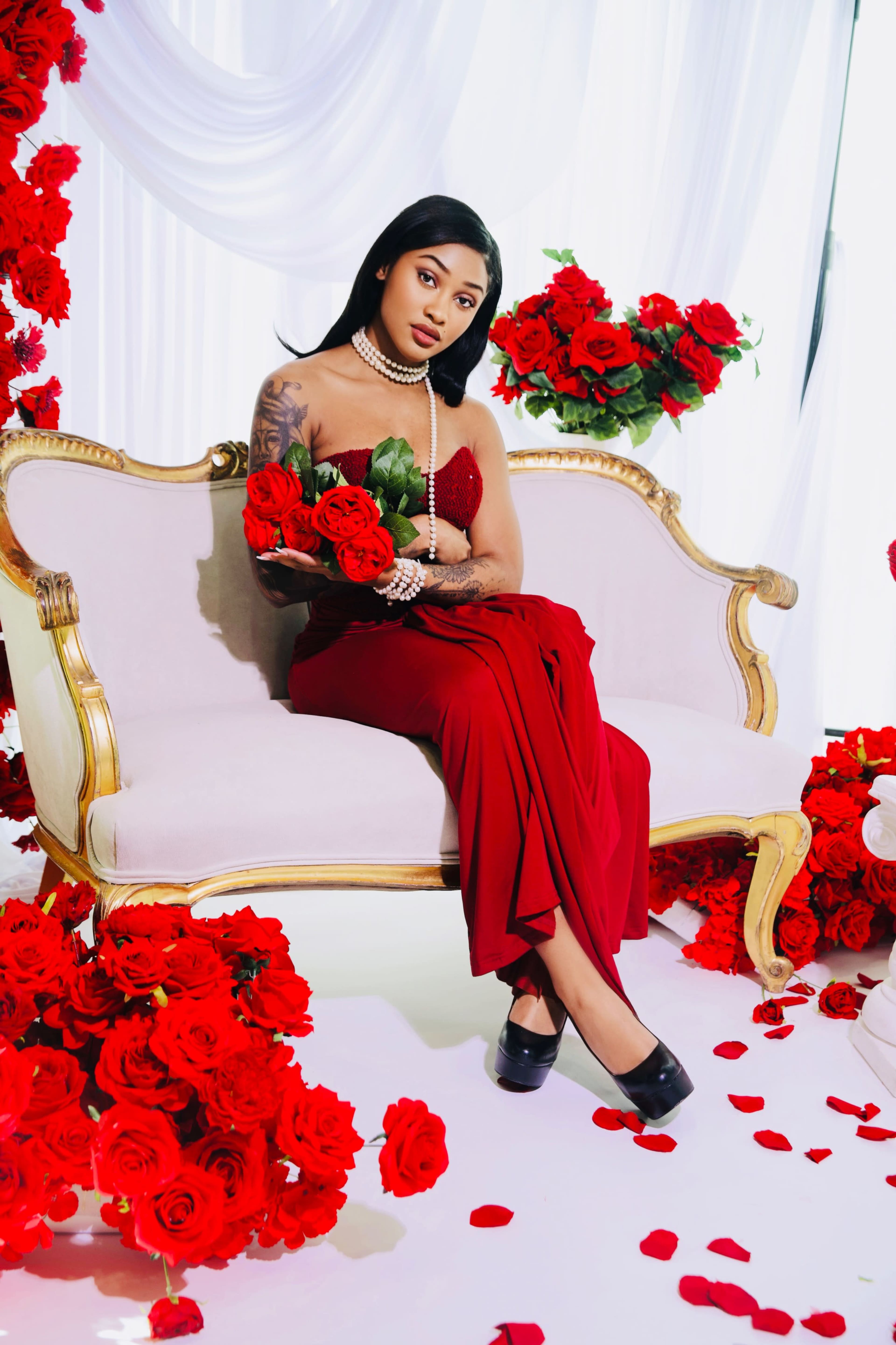 A woman in a red dress sits on a vintage couch adorned with artificial roses, holding a bouquet of red flowers amidst a backdrop of rose decorations.