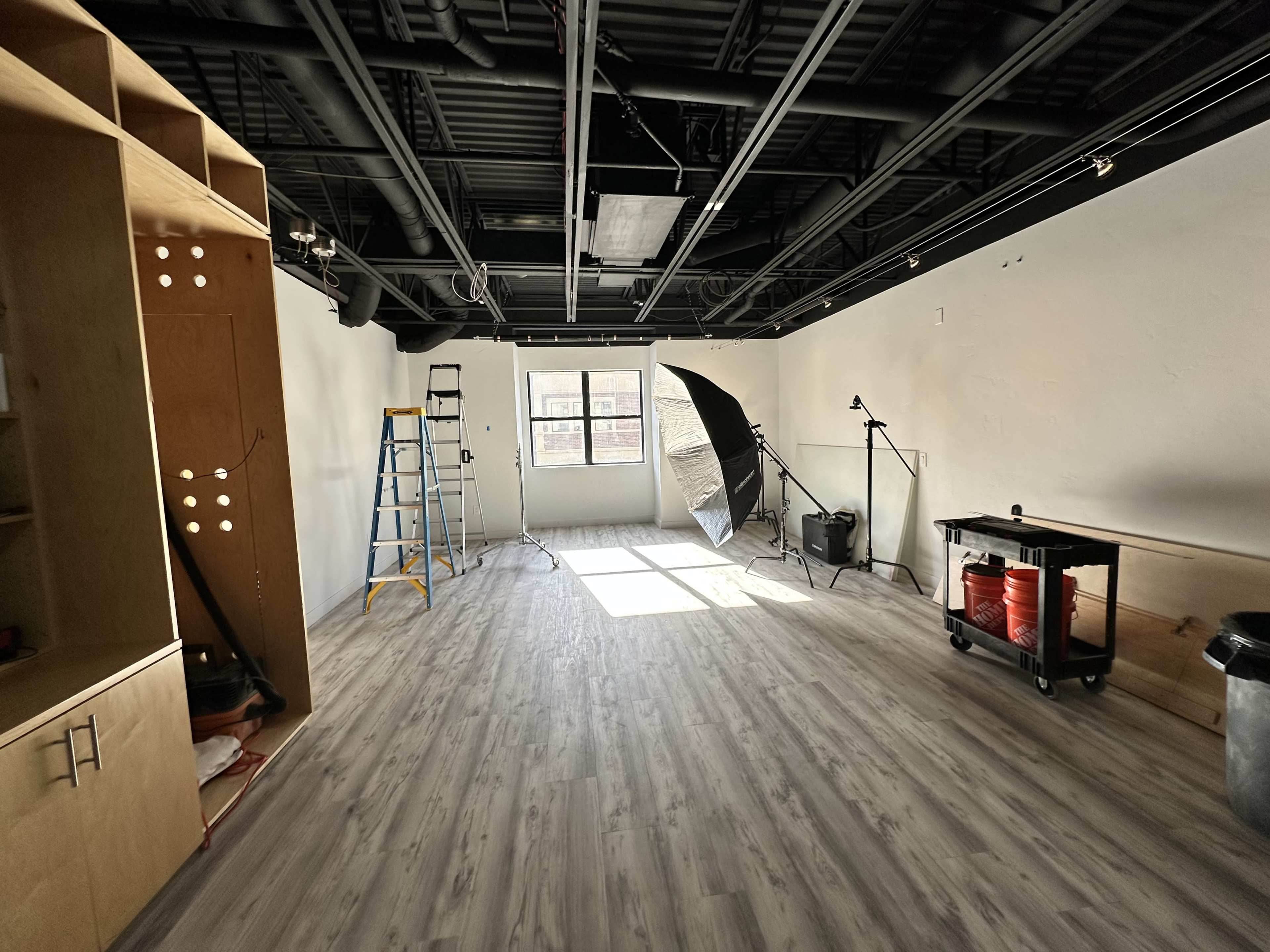 A spacious, empty room with wooden flooring, equipped with a ladder, light umbrella, and studio equipment.