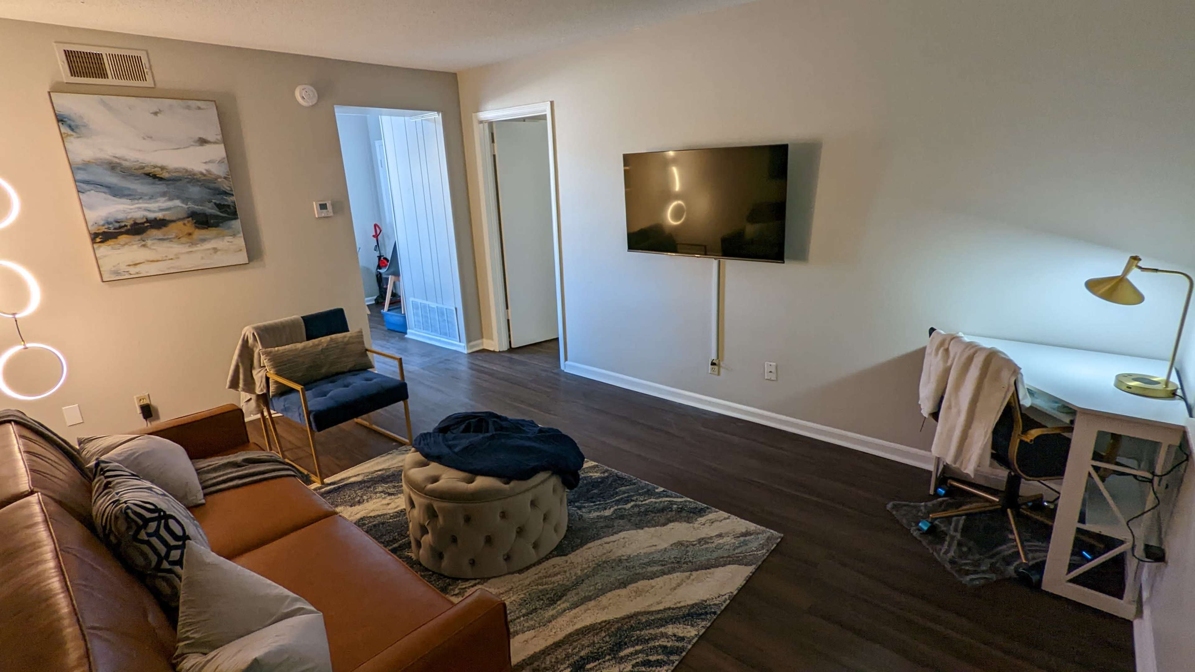 A living room features a brown leather couch, a round ottoman, a mounted TV, a small desk with a lamp, and a painting on the wall.