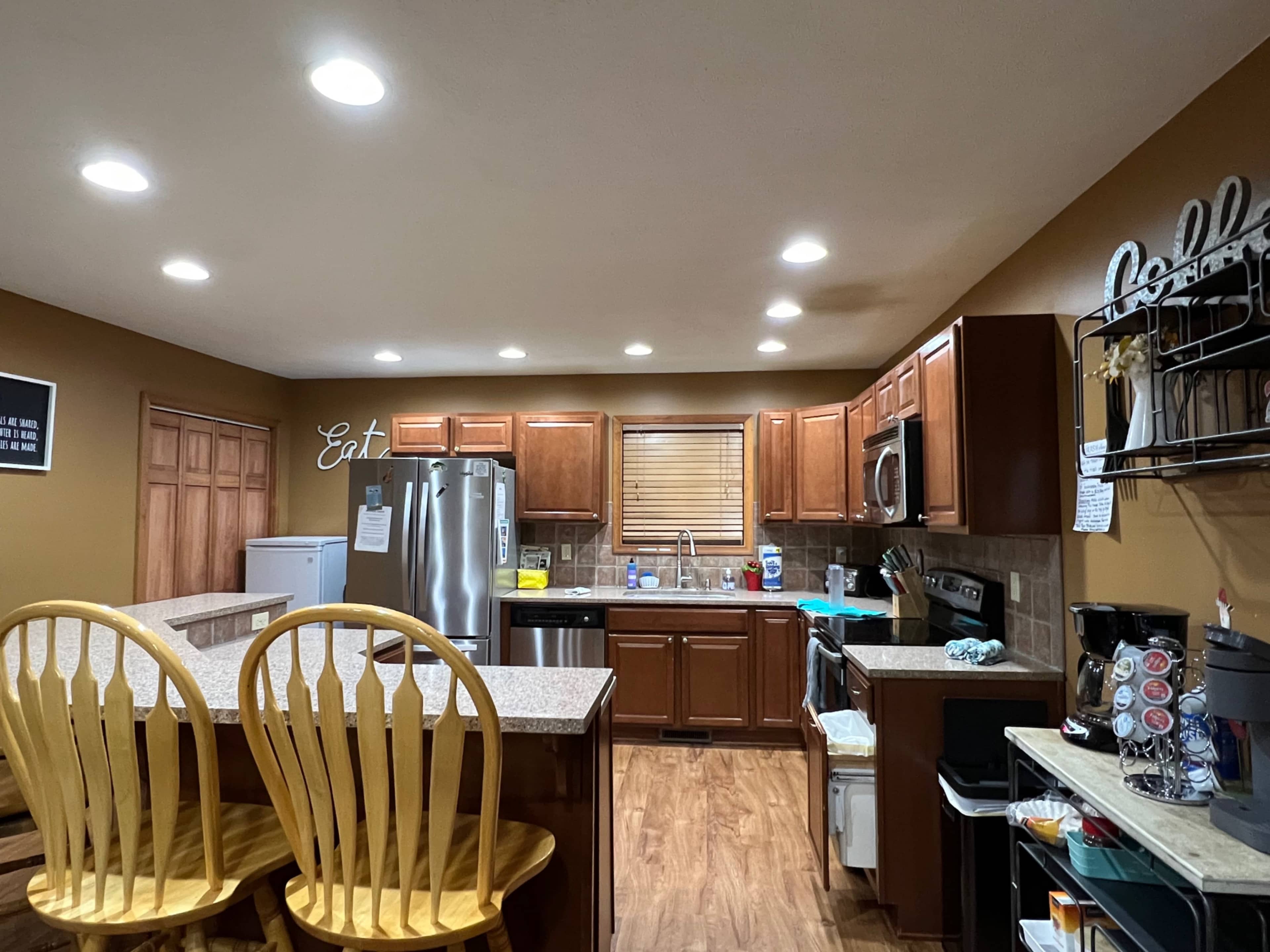 A kitchen with wooden cabinets, a refrigerator, a stove, and a countertop with yellow bar chairs, illuminated by recessed ceiling lights.