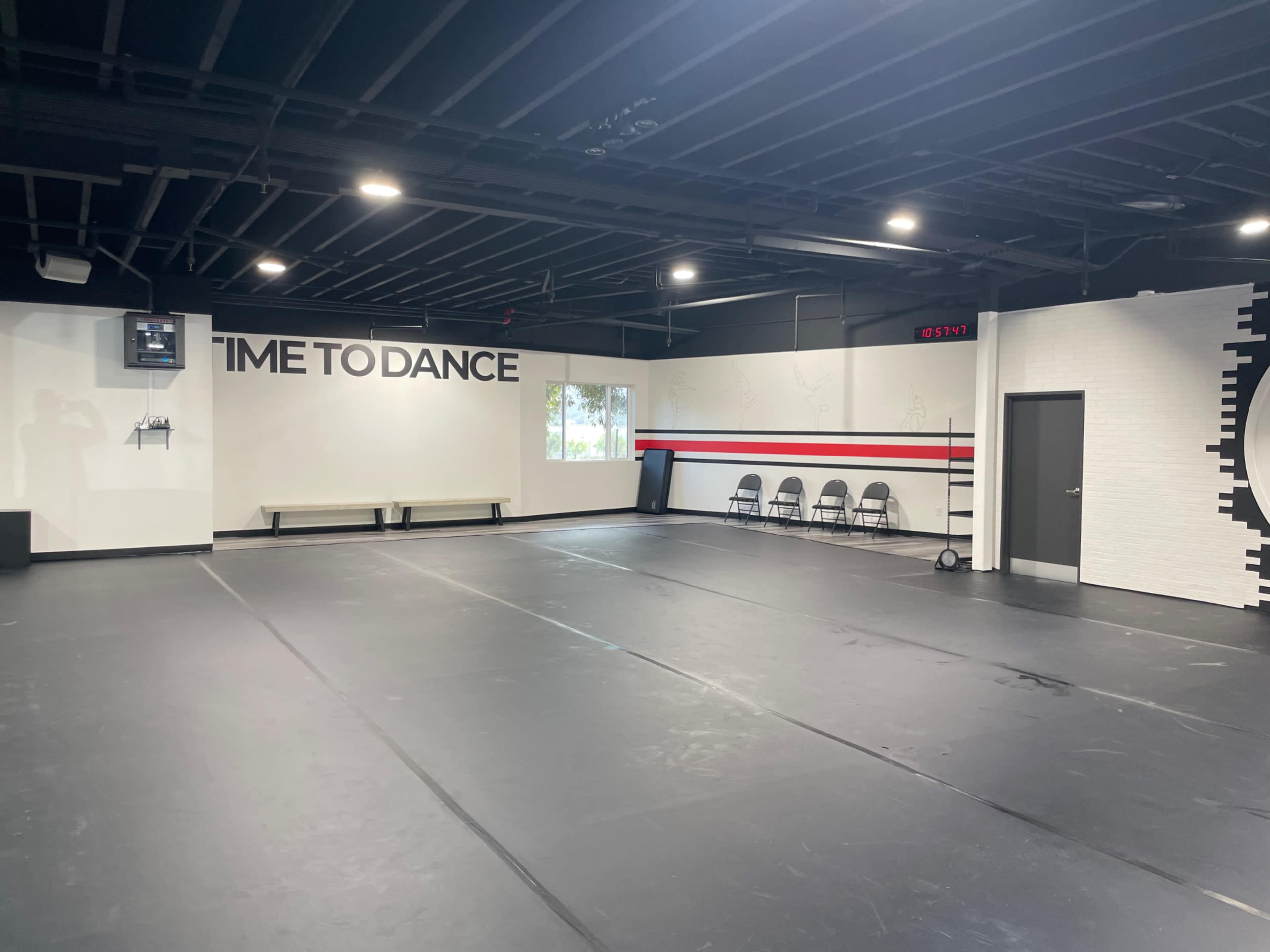An empty dance studio with a black floor, white walls featuring a "TIME TO DANCE" sign, and a row of chairs along one side.