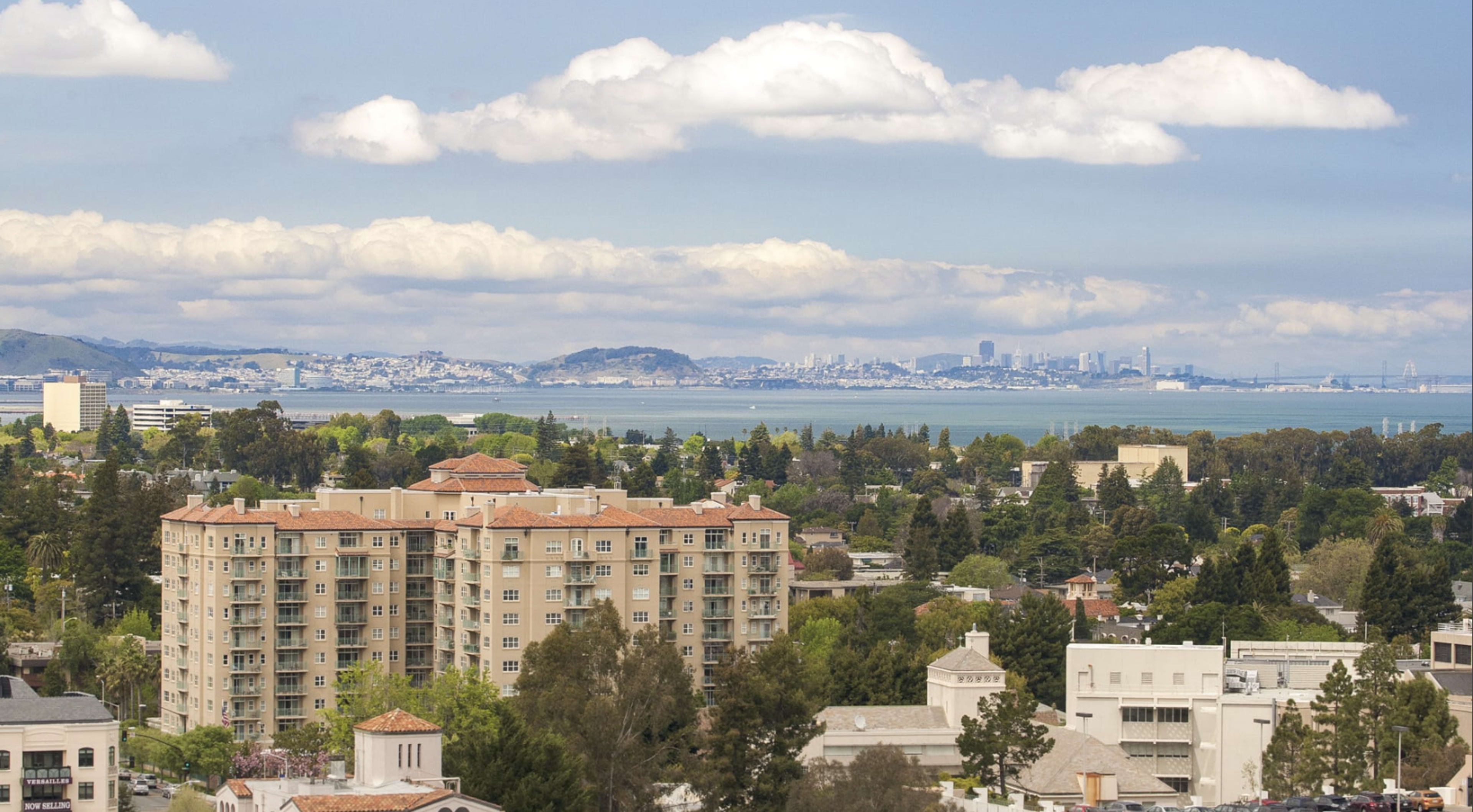 The image shows a suburban area with mid-rise buildings in the foreground and a distant city skyline across a body of water under a partly cloudy sky.