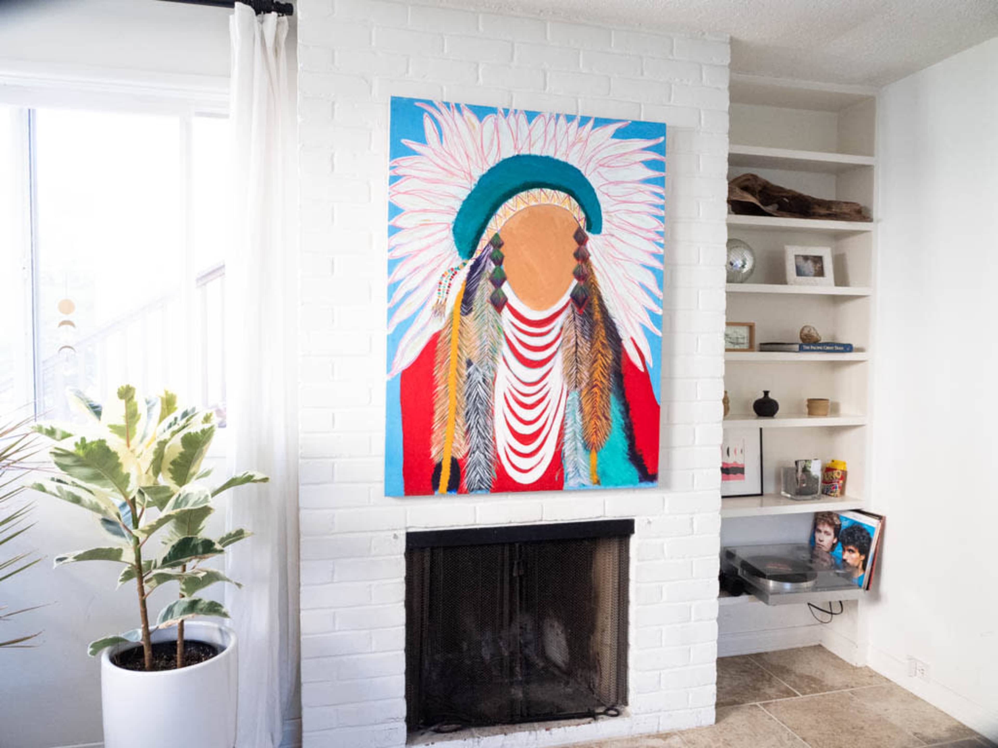 The image shows a living room with a white brick fireplace, a colorful painting of a figure with a headdress above it, and a plant in a pot near the window.