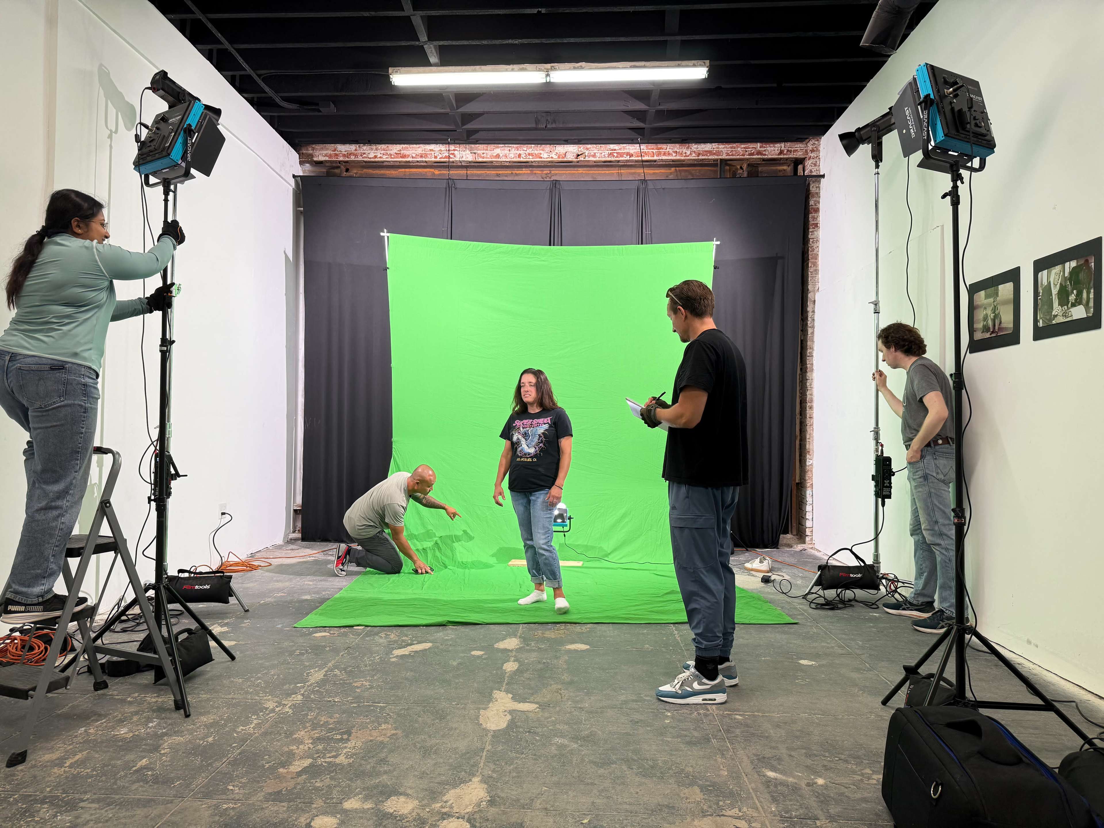 A group of six people is working in a studio setting with a green screen backdrop, where one person stands in front, while others adjust lighting and equipment.