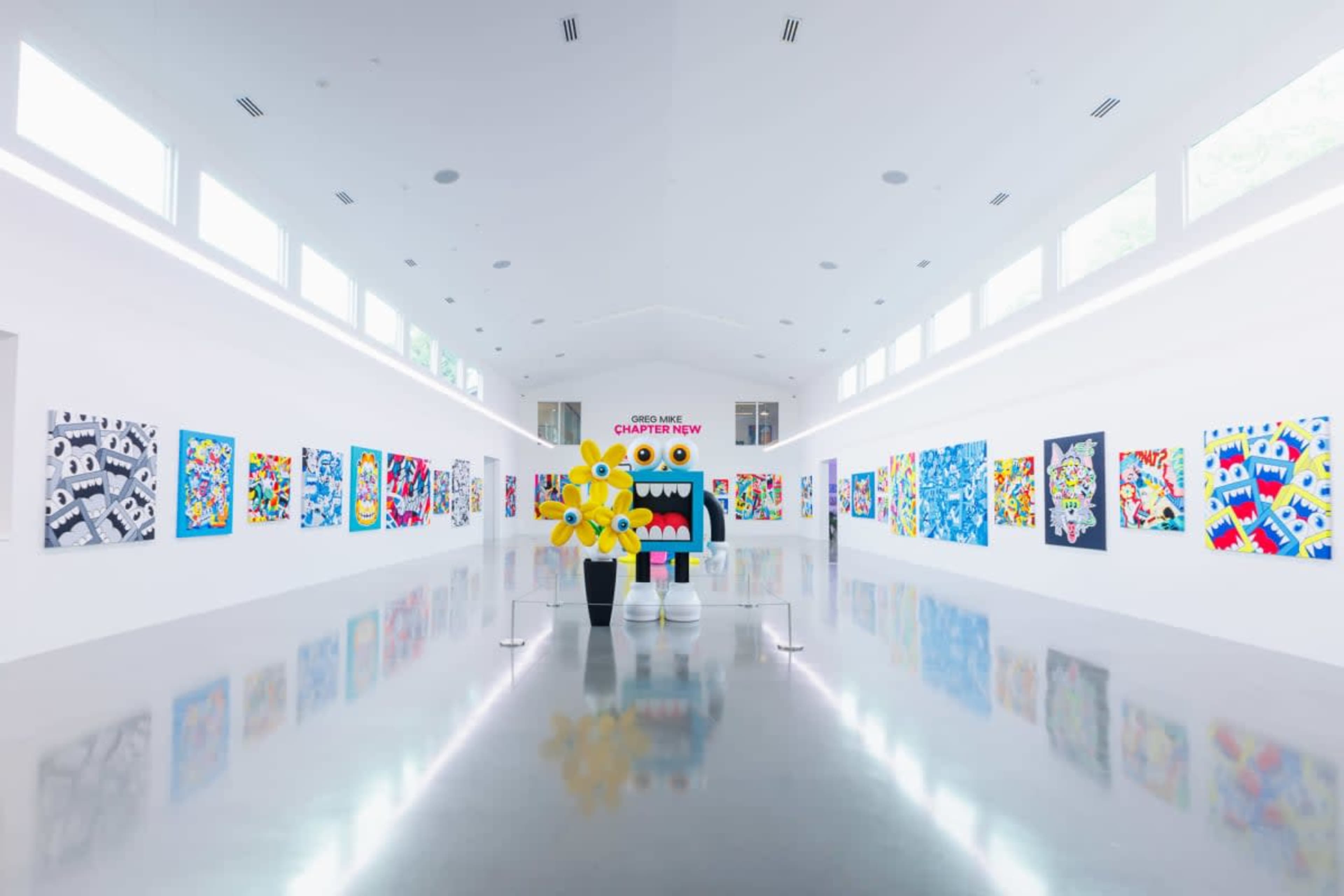 The image shows a modern art gallery with colorful artworks displayed on white walls and a large sculpture in the center.