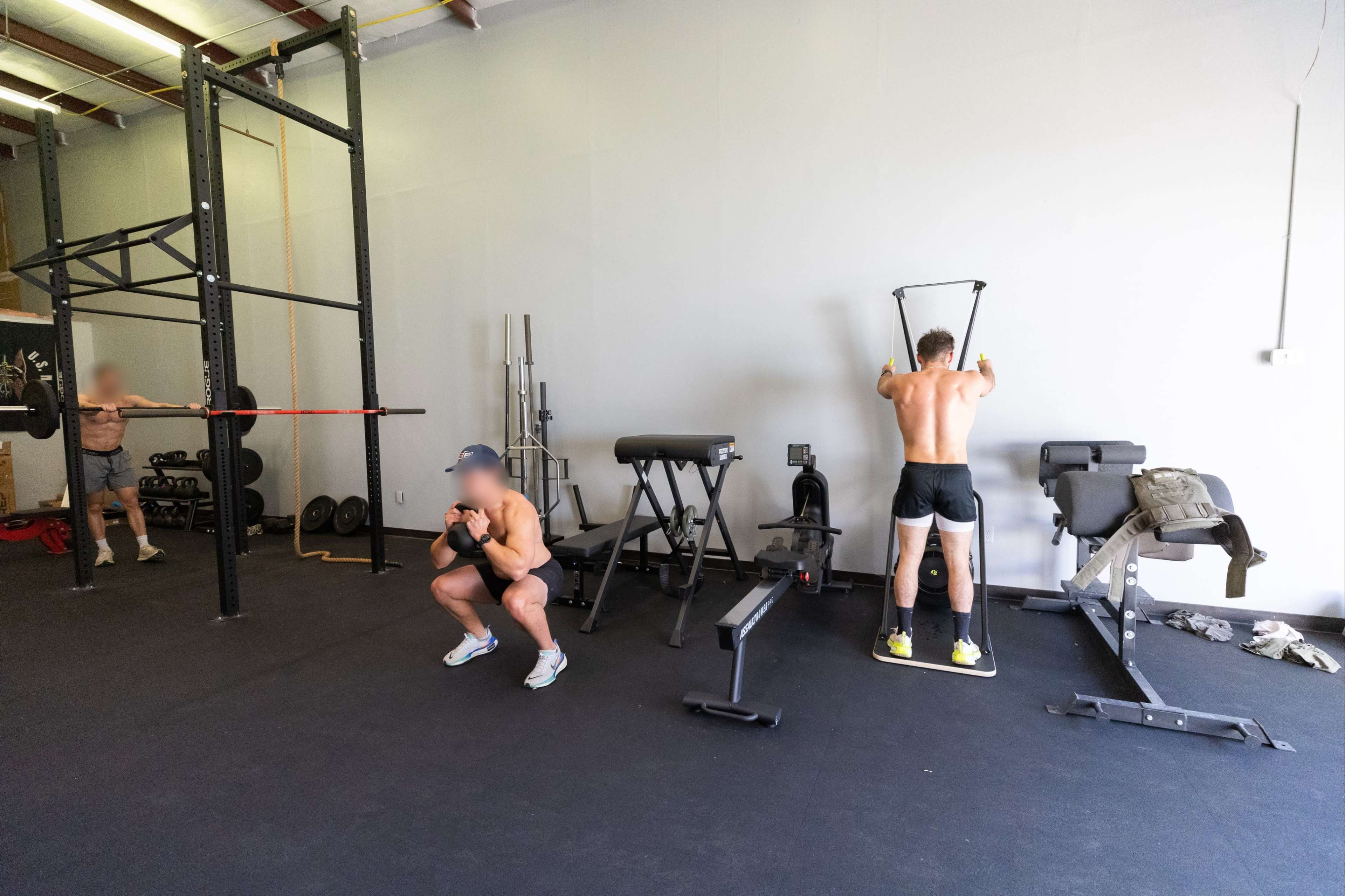Two men are exercising in a gym, with one performing squats while the other uses a resistance machine.