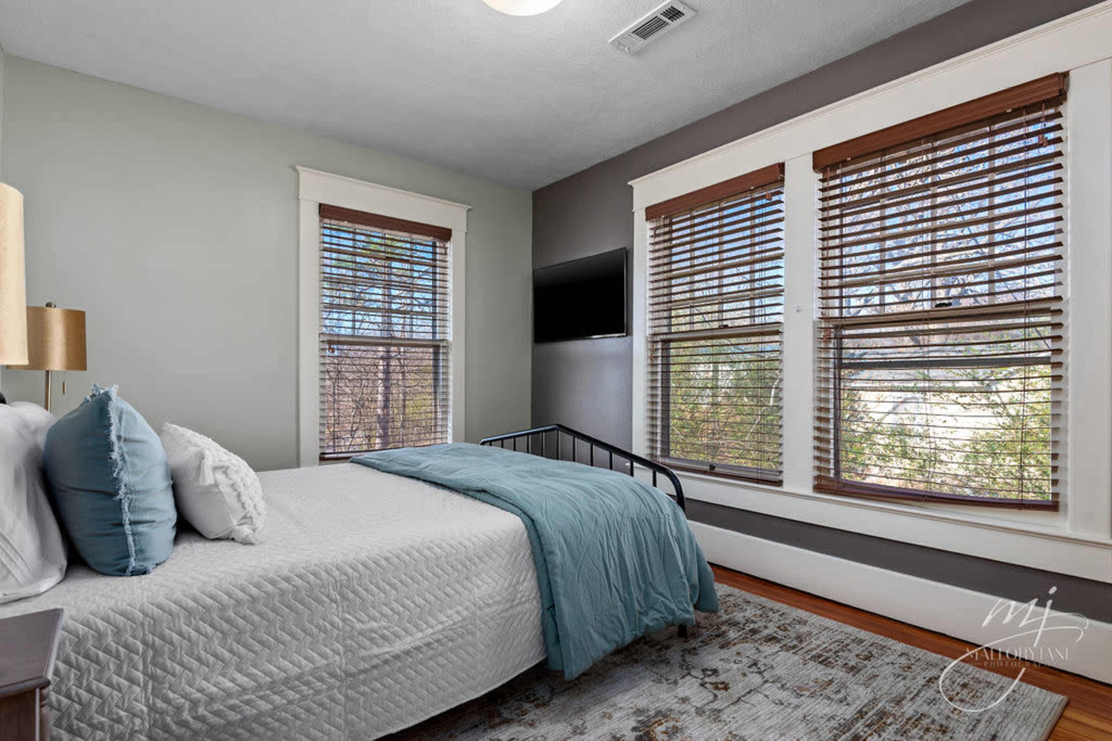 A bedroom features a bed with a light blue blanket, wooden blinds on the windows, and a flat-screen TV mounted on the wall.