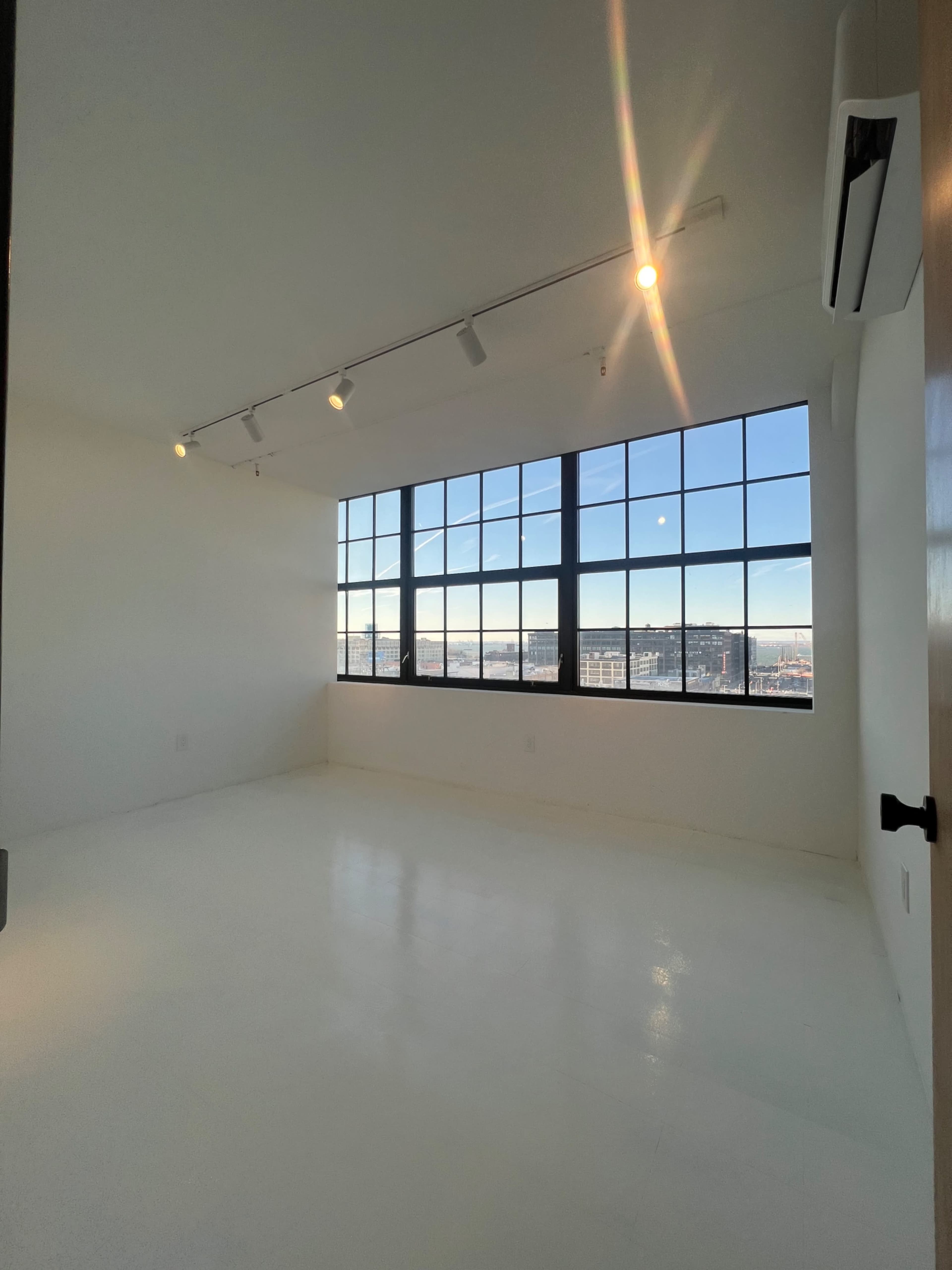 The image shows a spacious, brightly lit room with large windows offering a view of a cityscape.