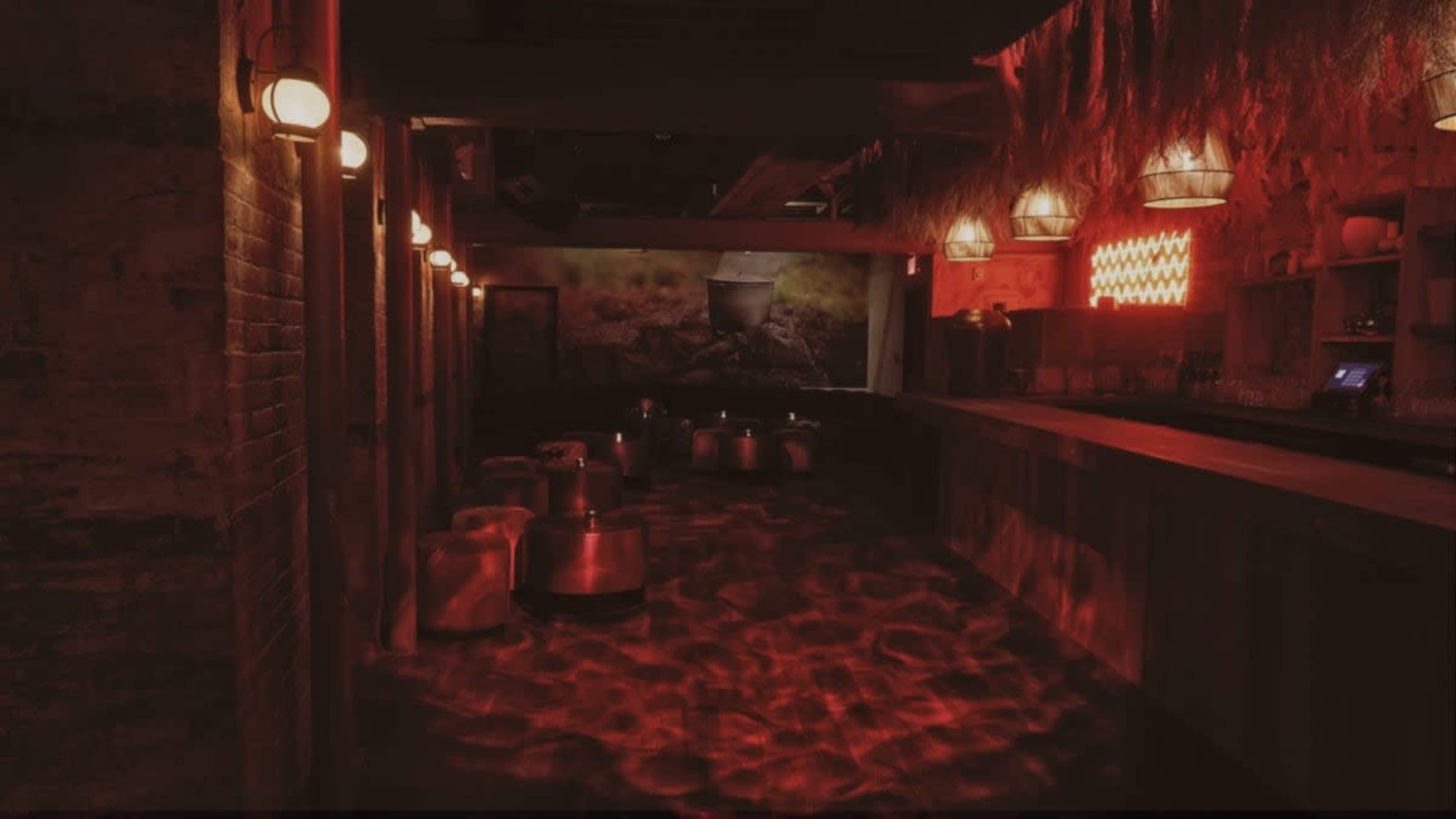 A dimly lit bar features a red-hued ambiance, with stools and tables arranged around a long counter.