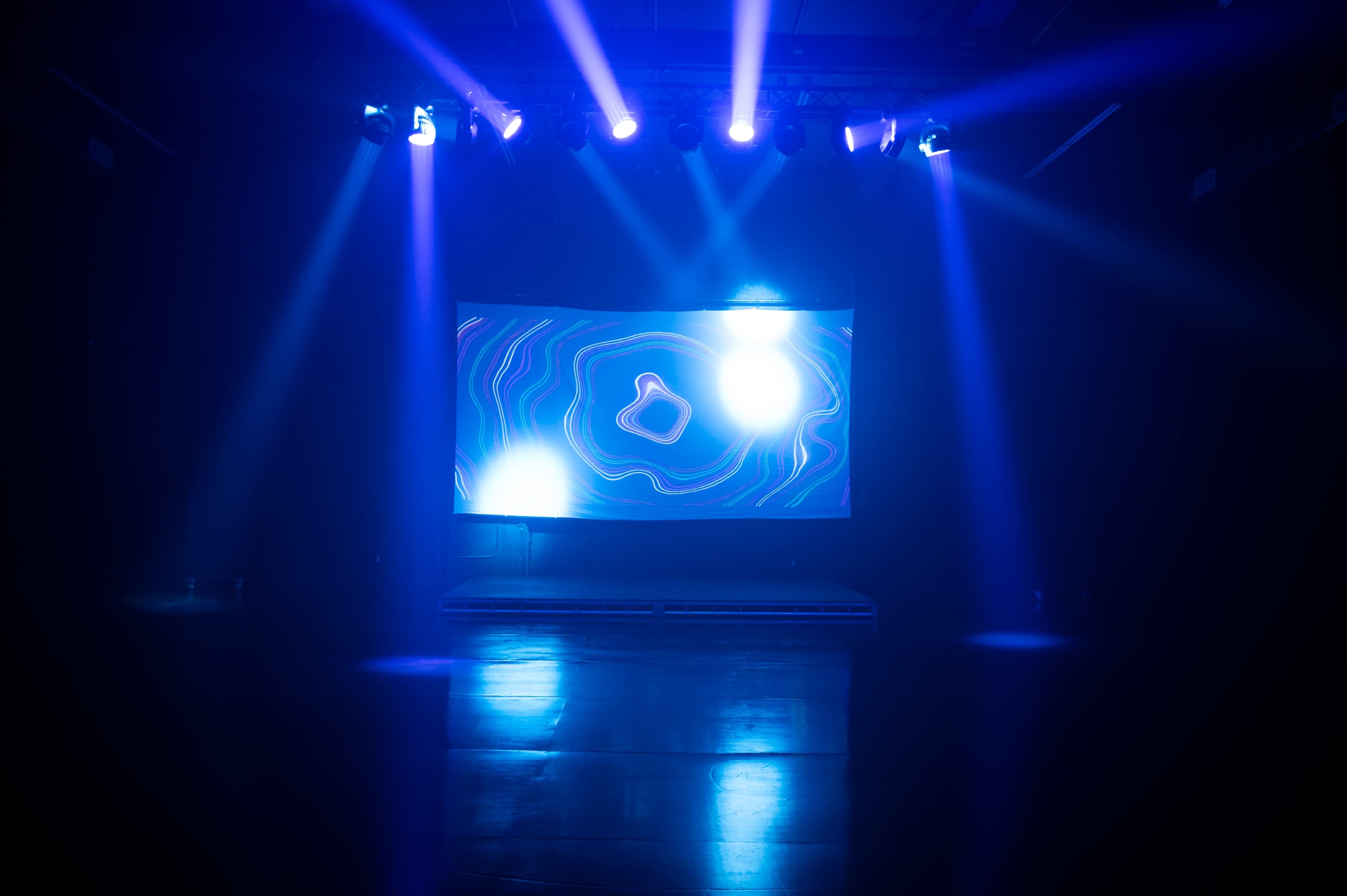 A stage with a large illuminated screen displaying dynamic graphics is surrounded by bright blue lights in a darkened room.