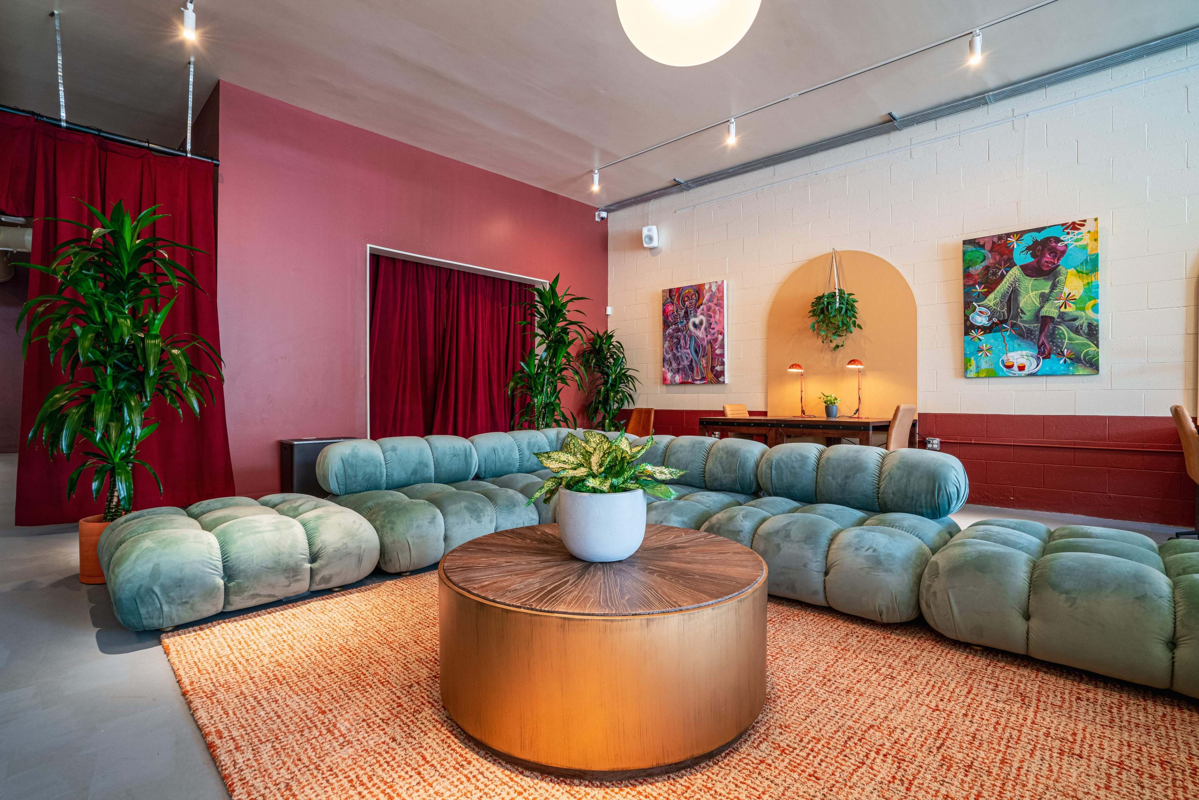 A modern lounge area featuring a large, round wooden table at the center, surrounded by plush, low seating and potted plants, with colorful artwork on the walls and red curtains in the background.