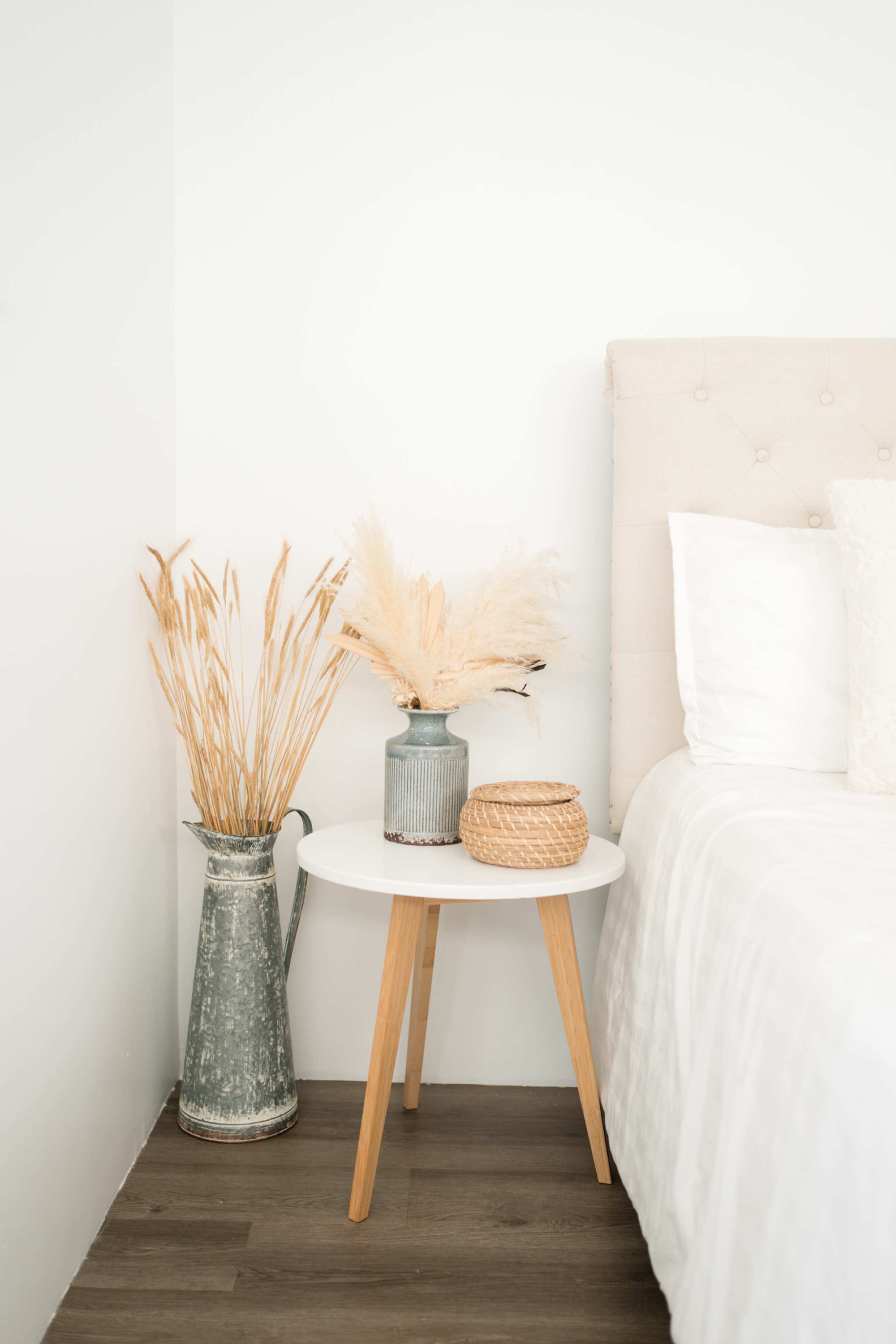A small white side table with wooden legs holds a gray vase with dried flowers and a woven basket, positioned next to a bed with white bedding against a plain wall.