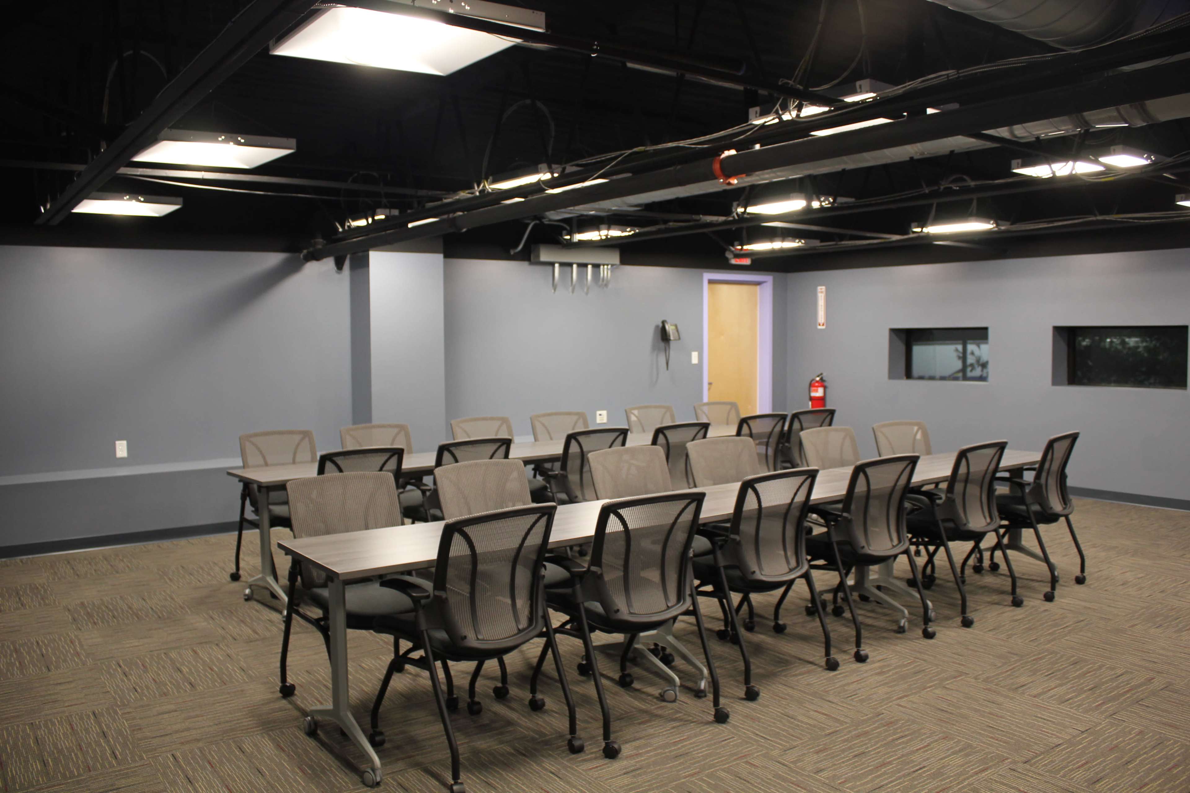 The image shows a classroom or meeting room with rows of movable tables and chairs arranged on a carpeted floor, under artificial lighting.