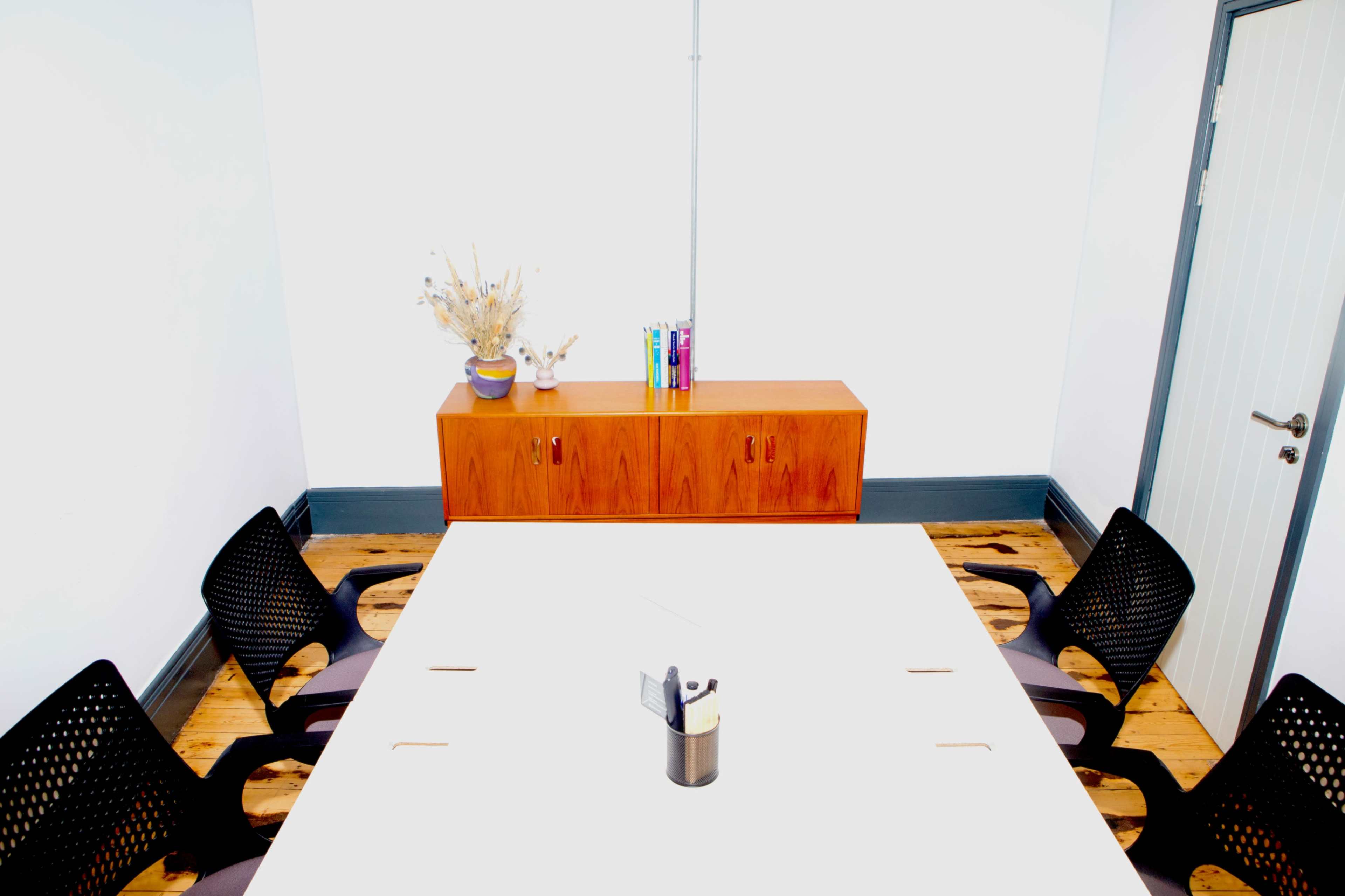 A minimalist conference room features a large white table surrounded by black chairs, with a wooden storage unit and decorative items against the wall.