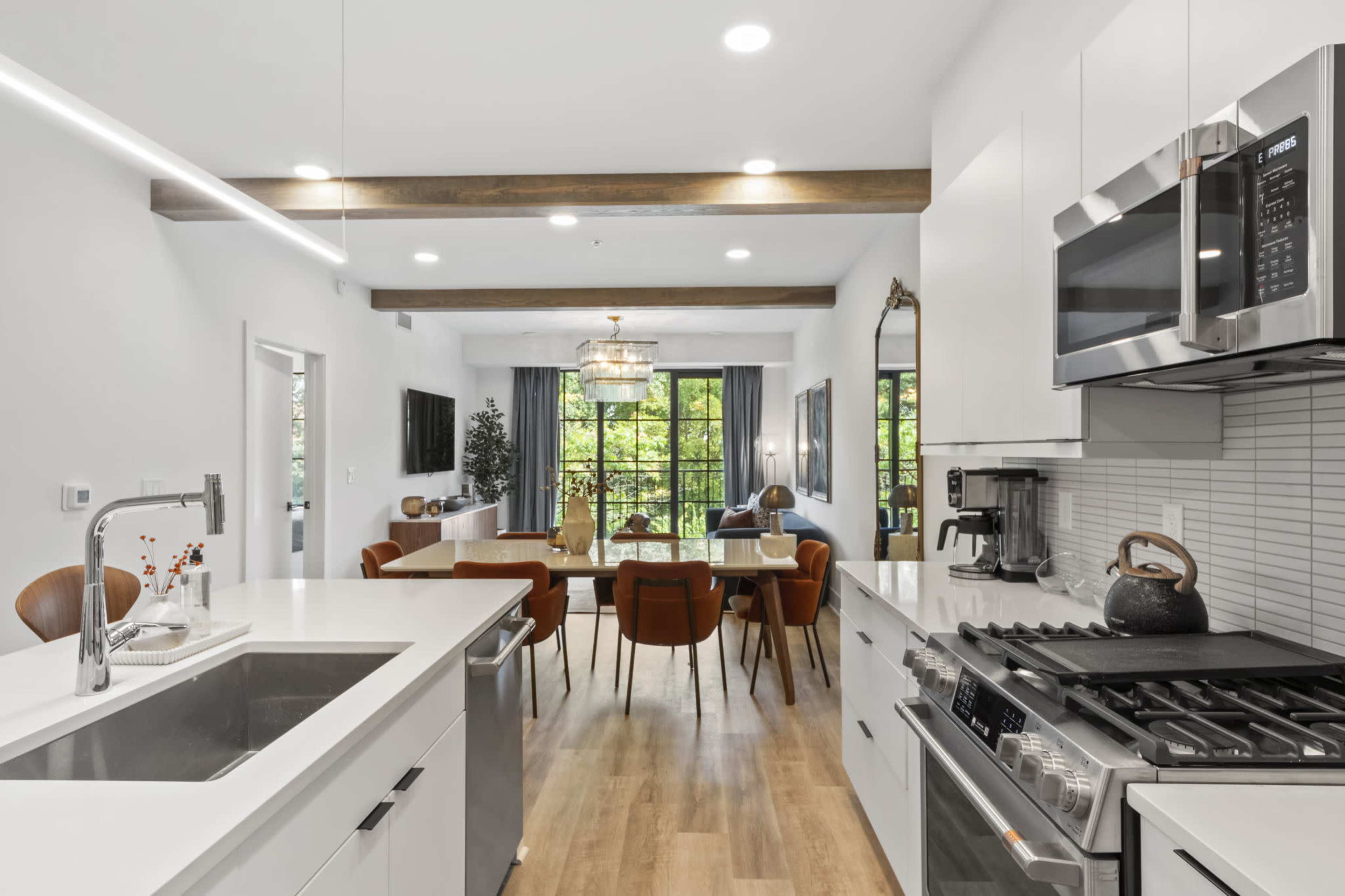 A modern kitchen with white cabinetry and stainless steel appliances opens into a dining area featuring a table surrounded by brown chairs and large windows overlooking greenery.