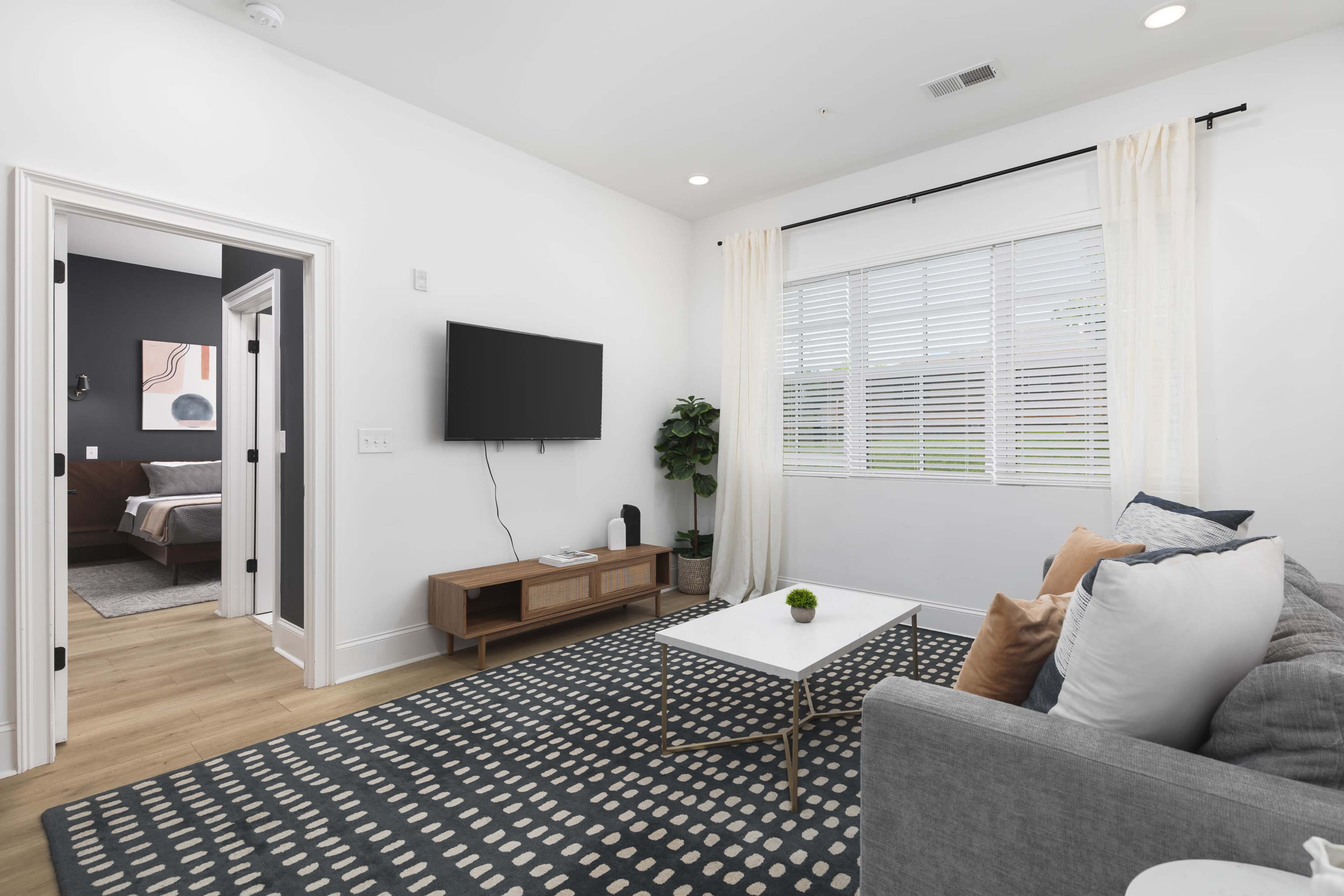 A modern living room features a gray sofa, a coffee table, and a television mounted on the wall, with a window dressed in curtains allowing natural light.