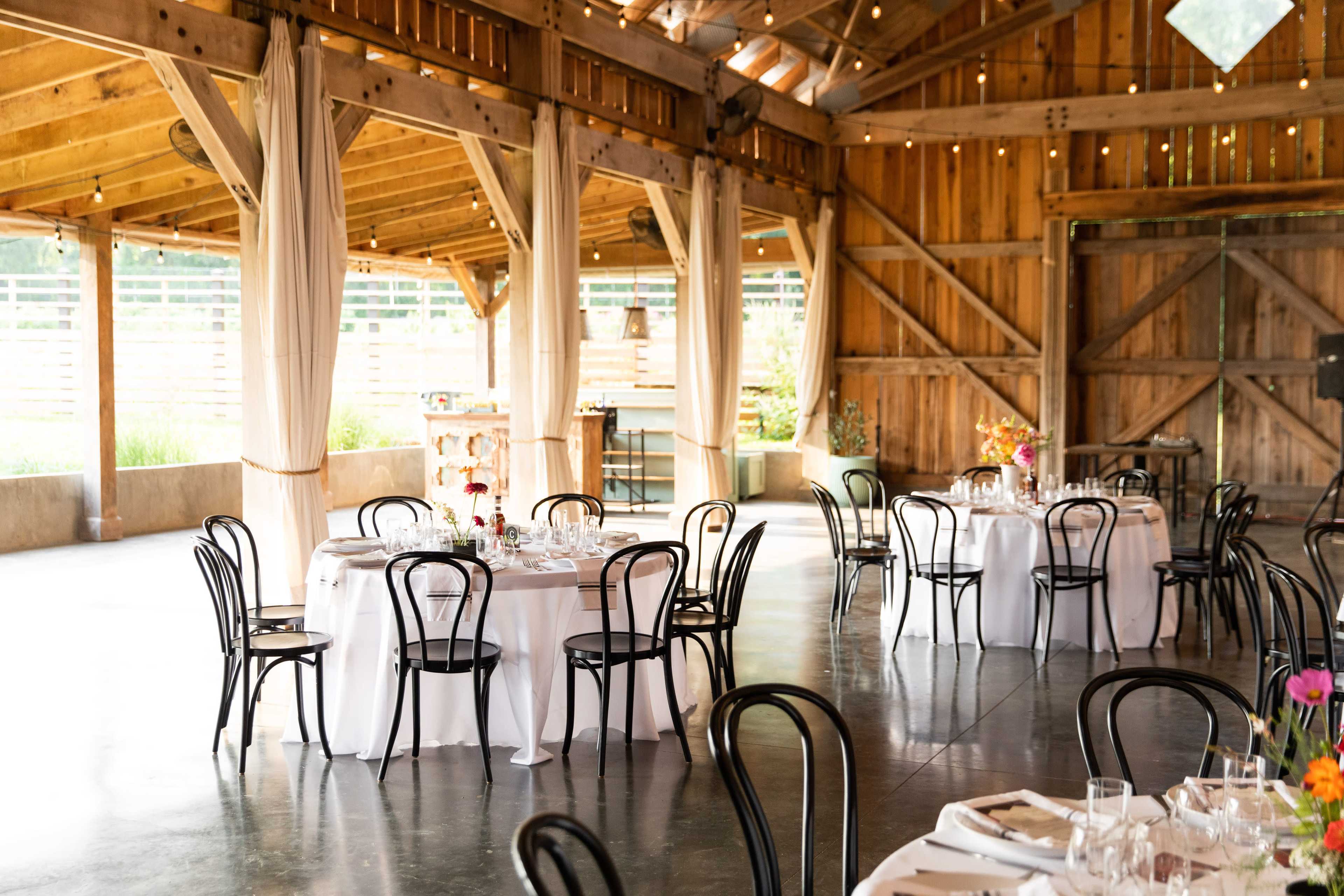 The image shows a rustic event space with wooden beams, tables set for dining, and decorative floral arrangements.