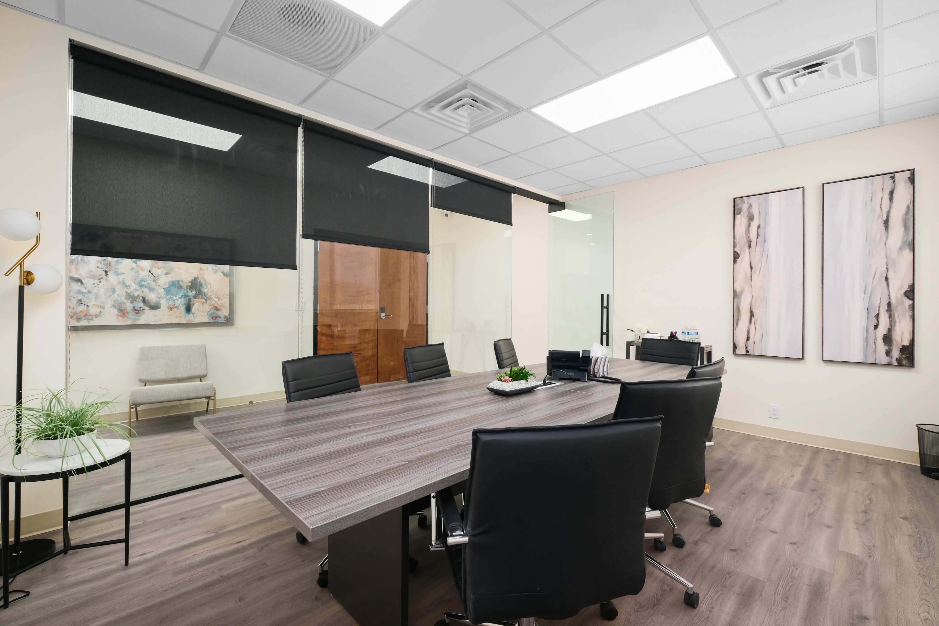 A modern office meeting room with a large table surrounded by black chairs, glass walls, and abstract artwork on the adjacent walls.