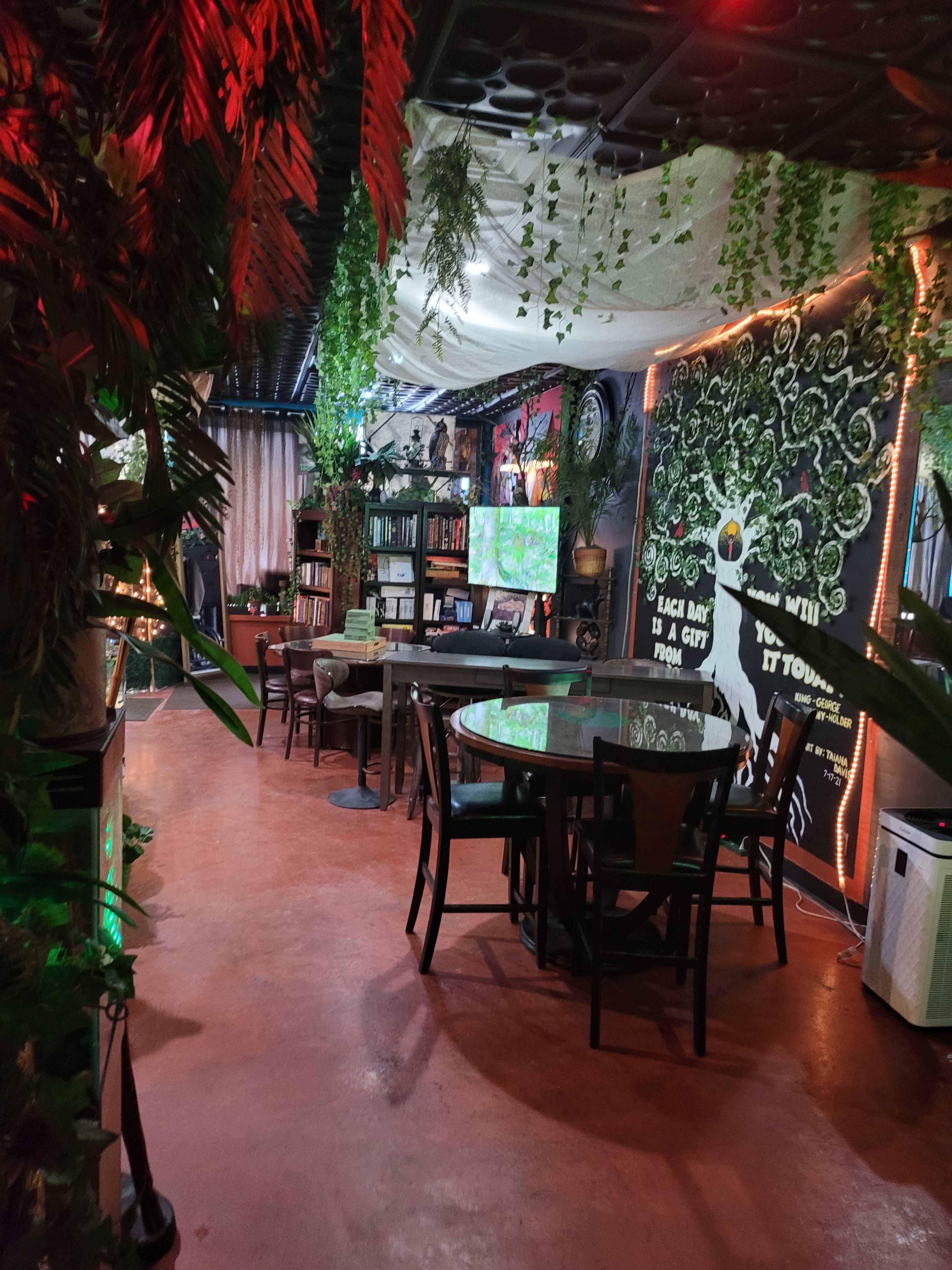 A brightly lit café with greenery, bookshelves, and a large wall mural.