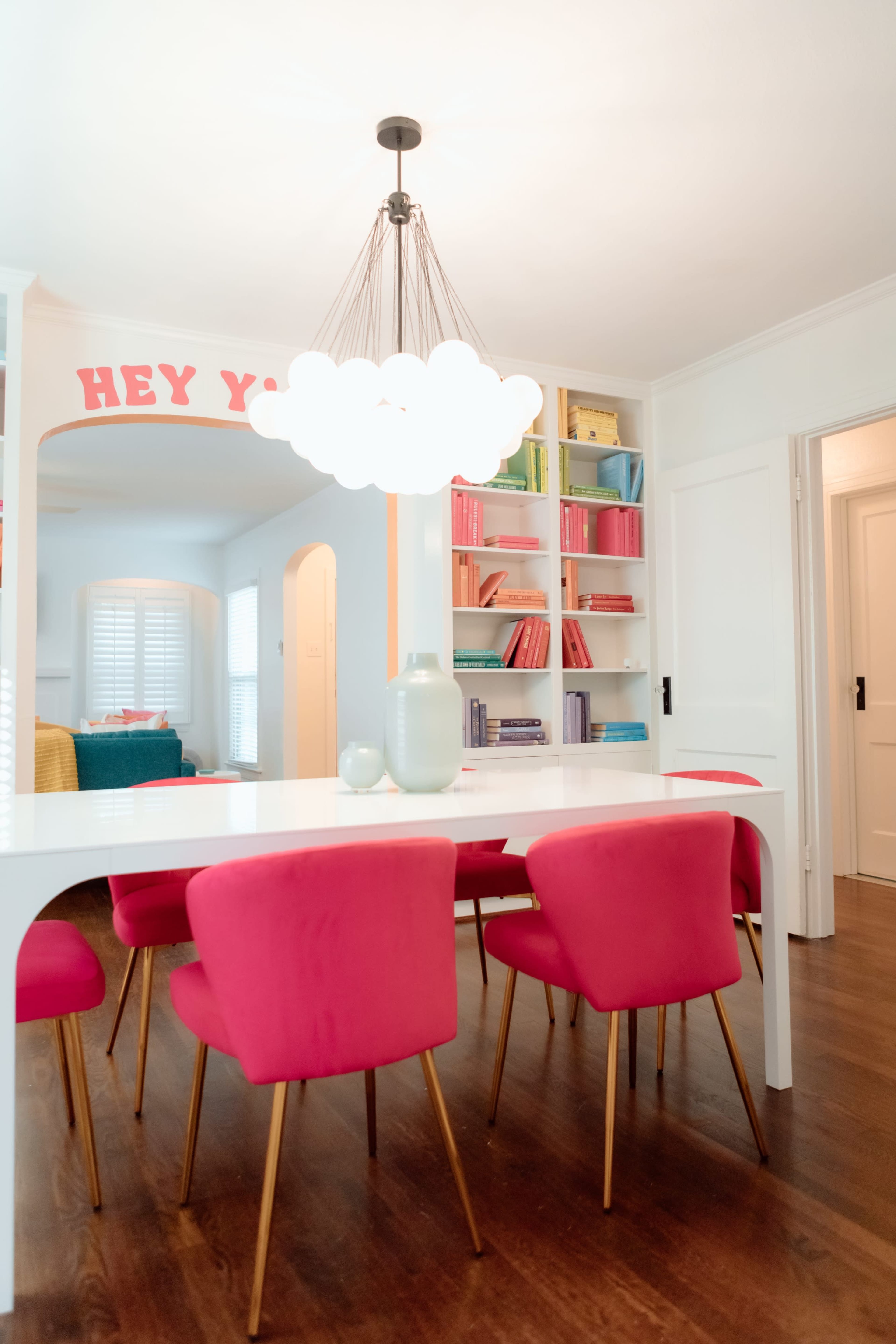 A brightly lit dining area features a white table surrounded by pink chairs and a colorful bookshelf in the background.
