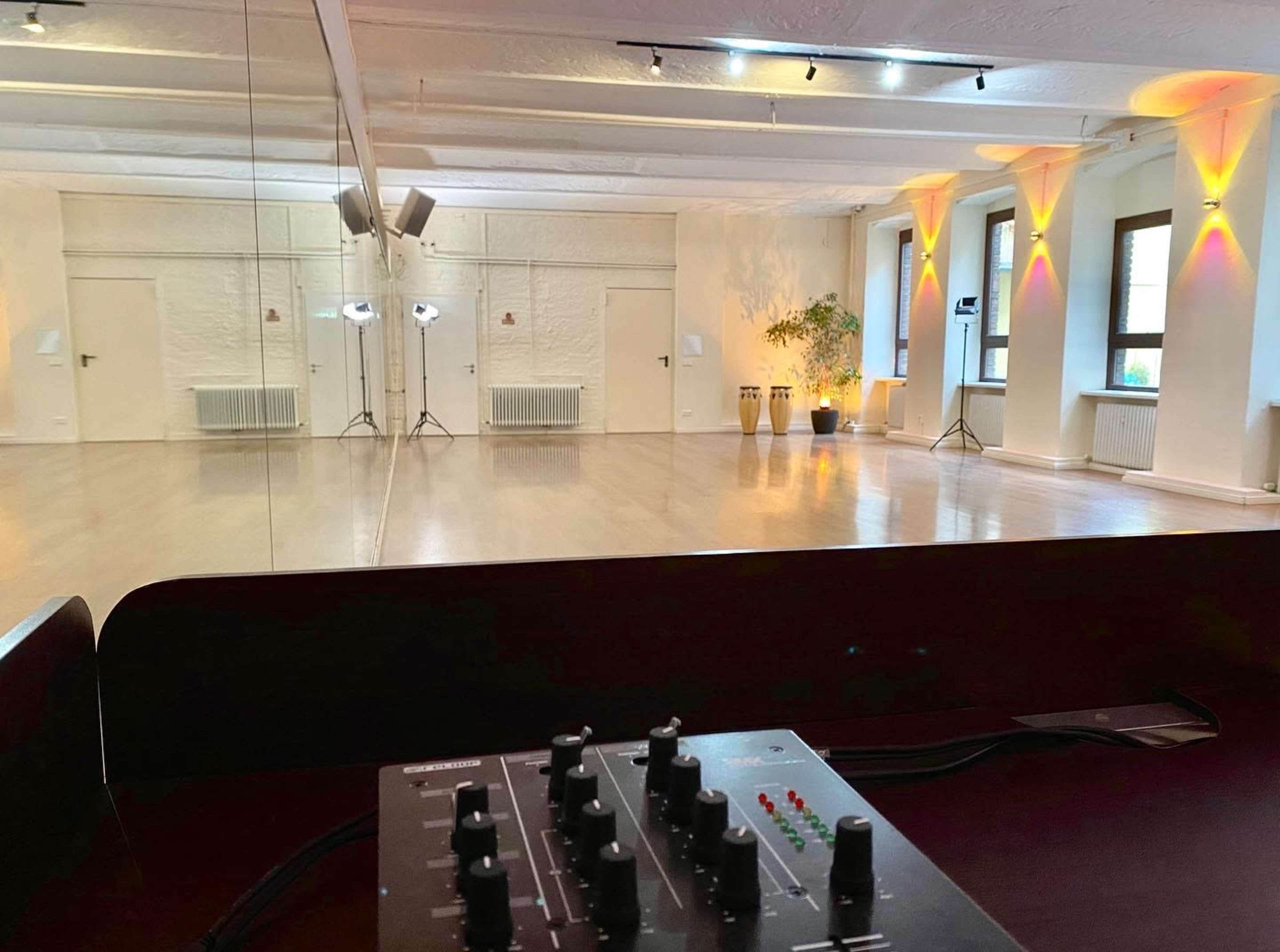 An empty studio space with wooden flooring, wall-mounted lighting, and sound equipment in the foreground.