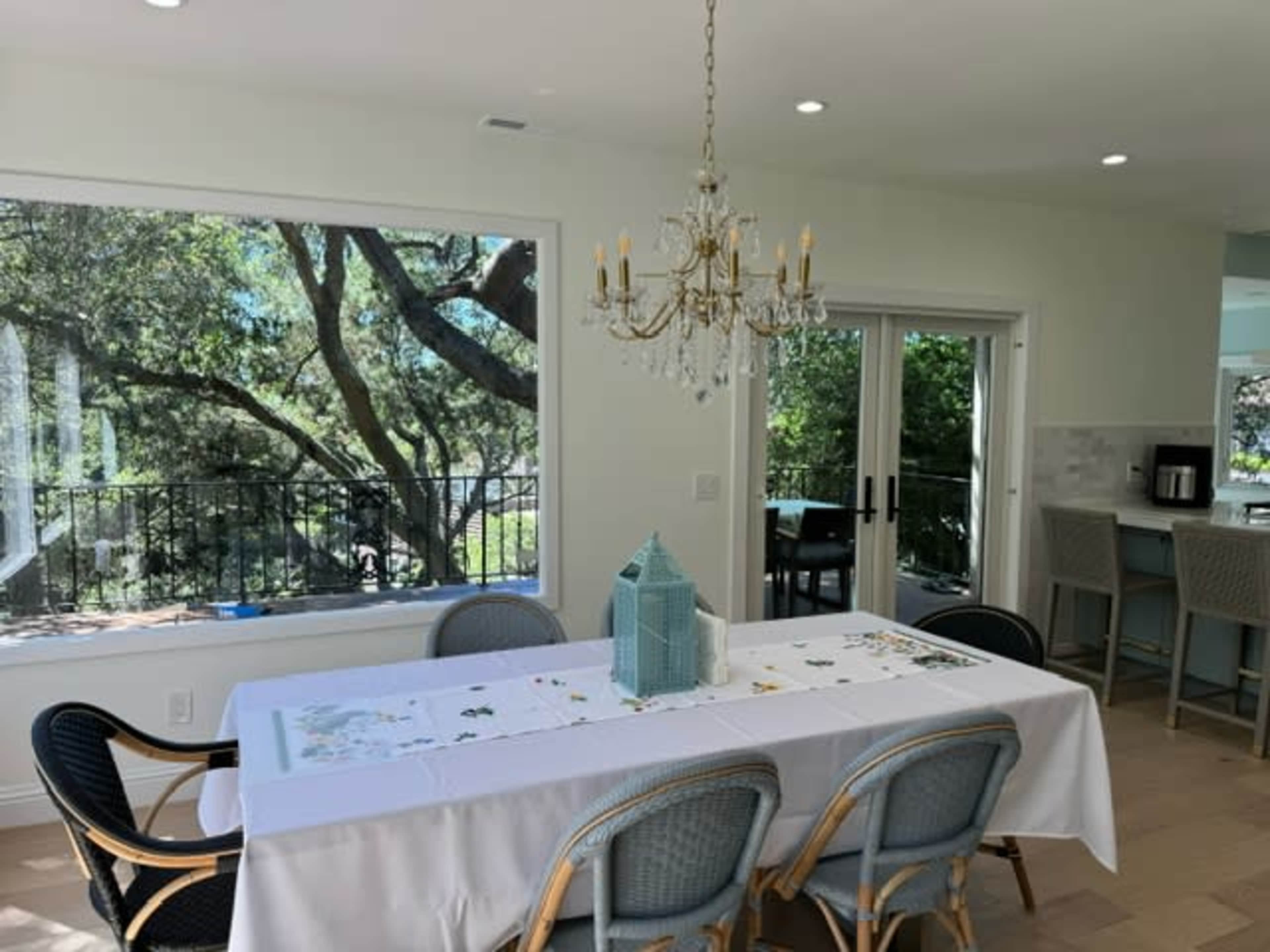 A dining area features a white tablecloth draped over a long table, surrounded by wicker chairs, with a chandelier above and large windows revealing a view of trees outside.