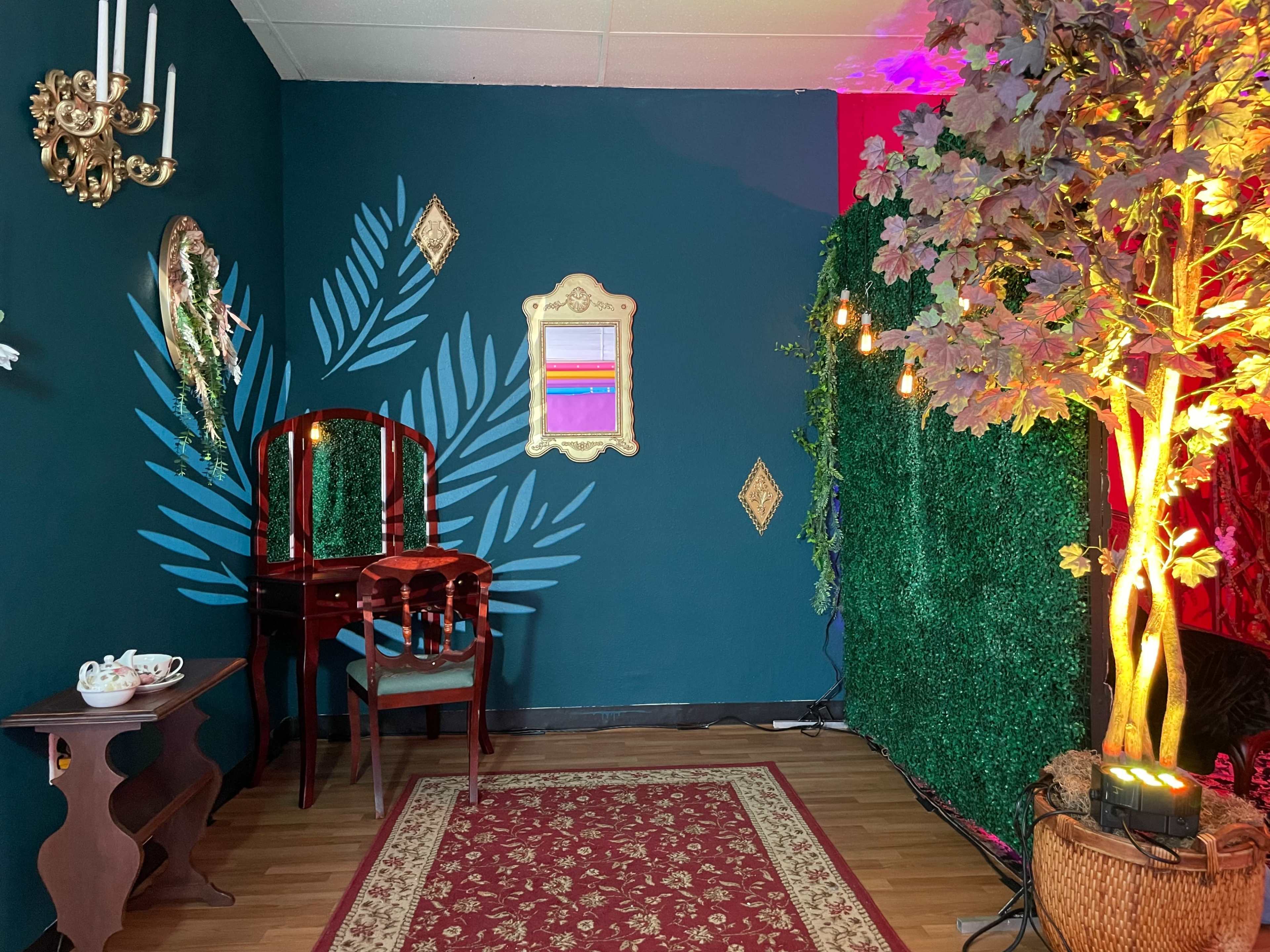 The scene features a compact room adorned with dark green walls, a wooden vanity with a chair, a decorative mirror, and illuminated foliage set against a backdrop of artificial greenery.