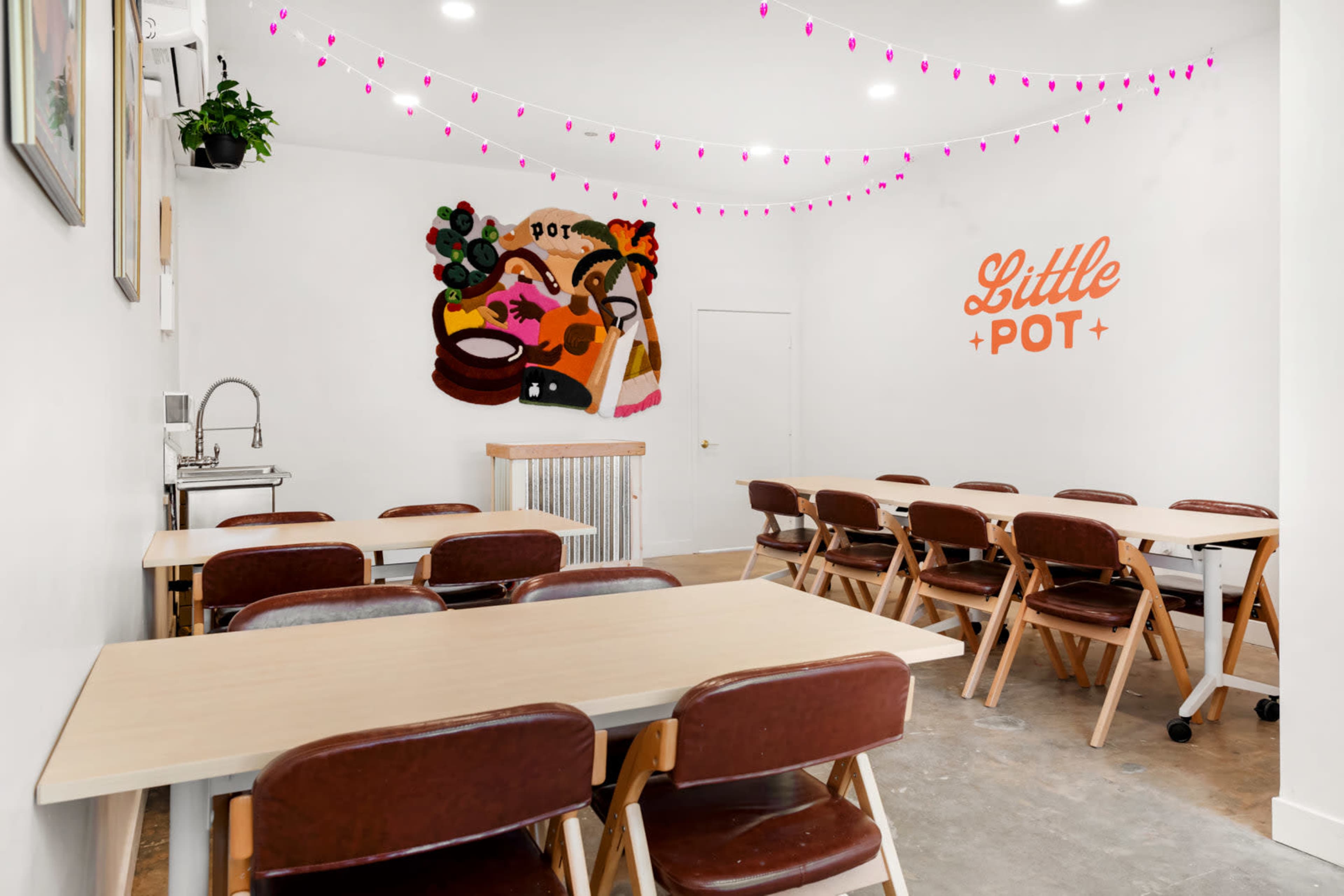 The image shows a bright, minimalist indoor space with several wooden tables and chairs arranged in rows, featuring a colorful mural on the wall.