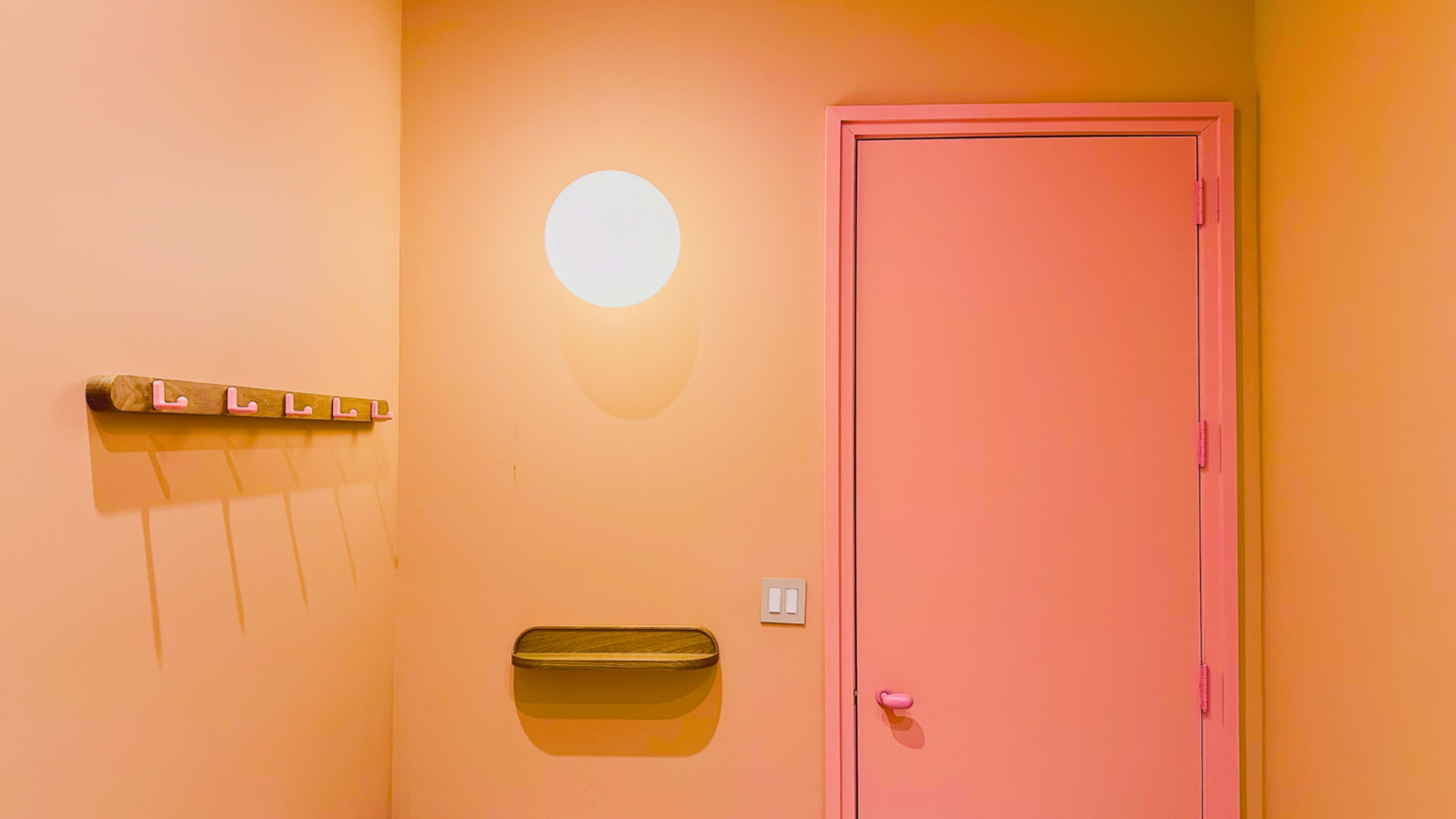 The image features a simple interior hallway with pastel orange walls, a round light fixture, a pink door, and a wooden shelf with wall hooks.