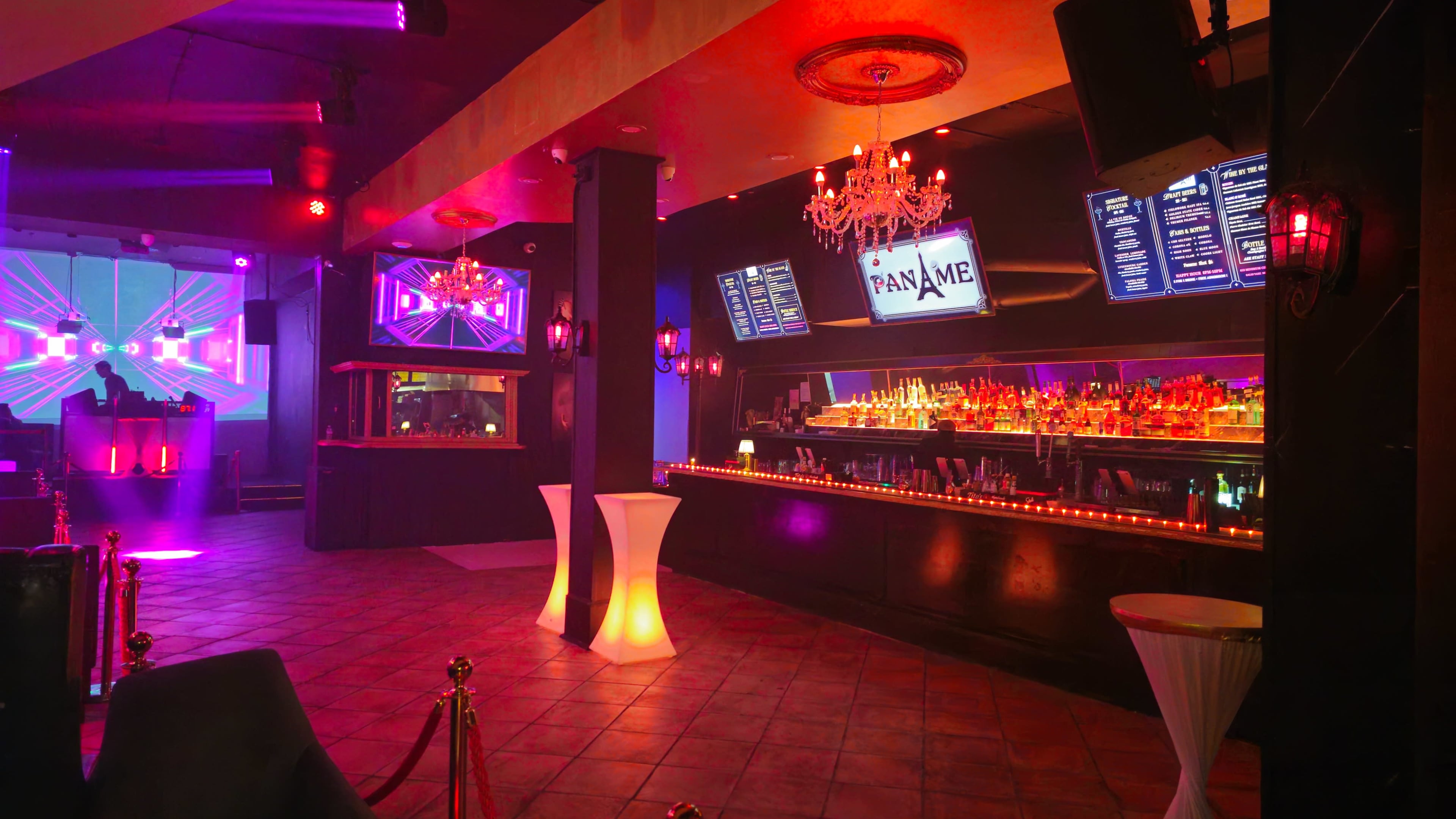 A dimly lit nightclub features a long bar illuminated by colored lights and multiple screens displaying visuals.