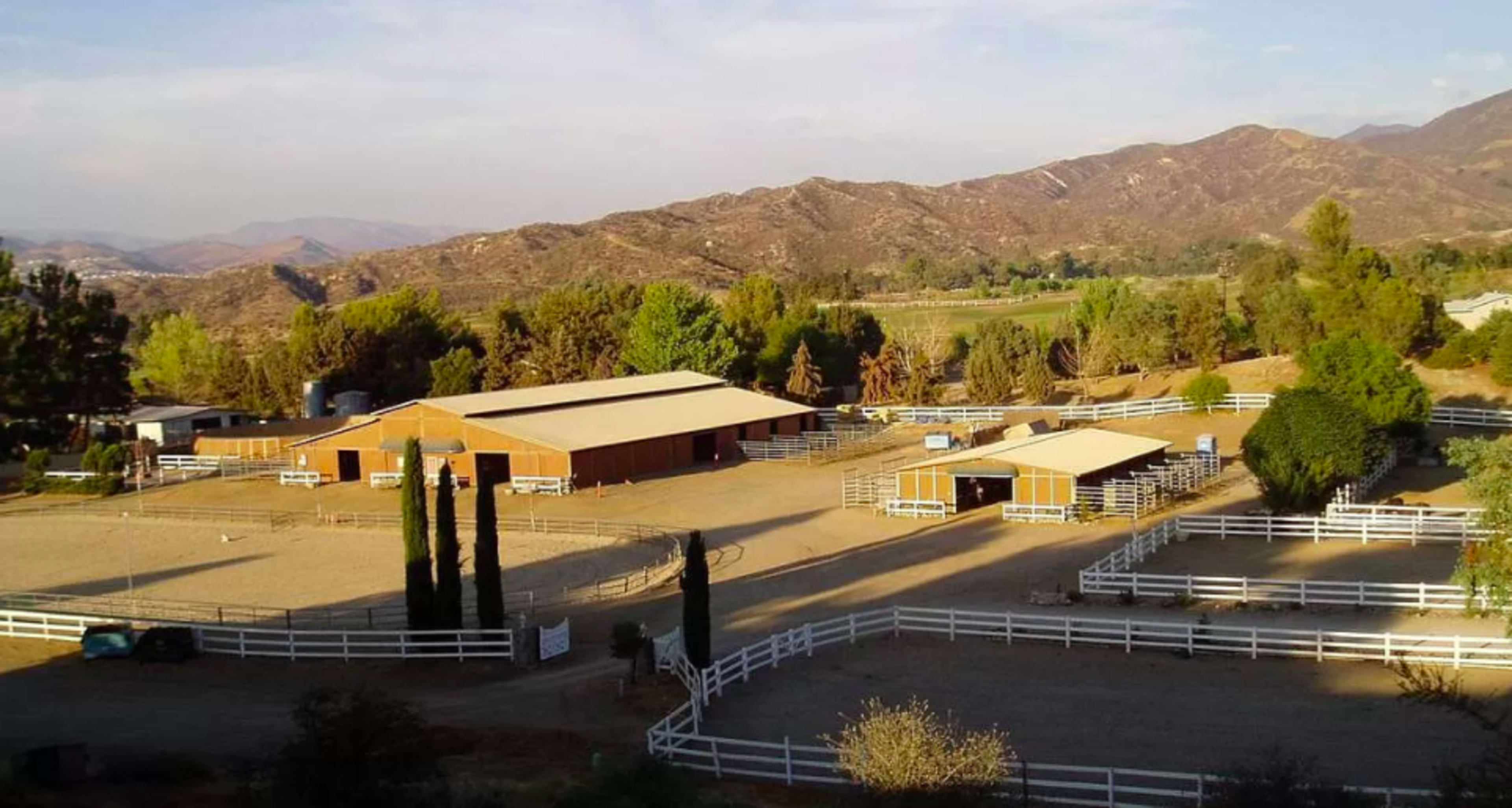 A sprawling equestrian facility features several wooden barns and riding areas, surrounded by mountains and trees.