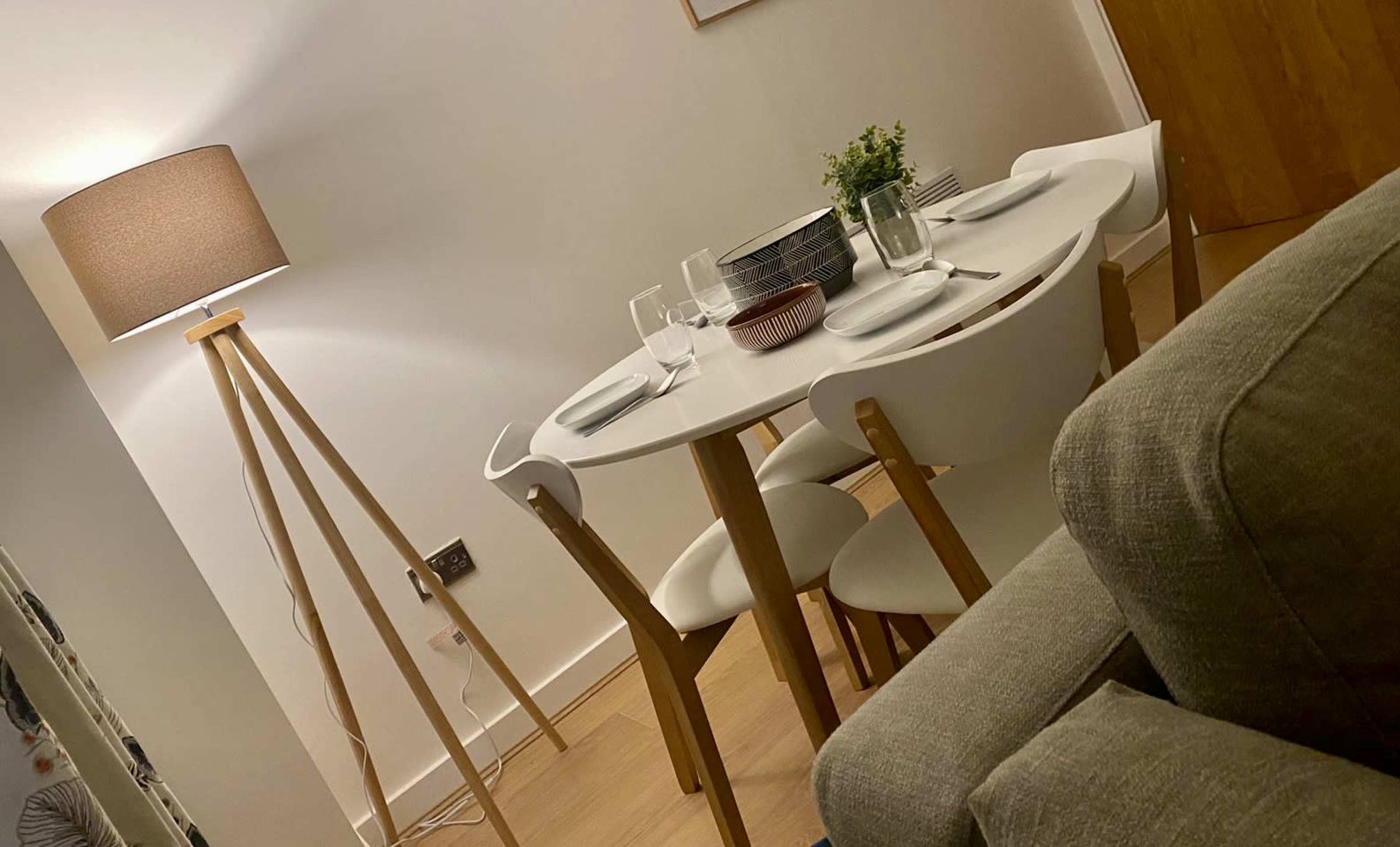 A modern dining room features a round table set for a meal, surrounded by white chairs, with a floor lamp and a couch nearby.