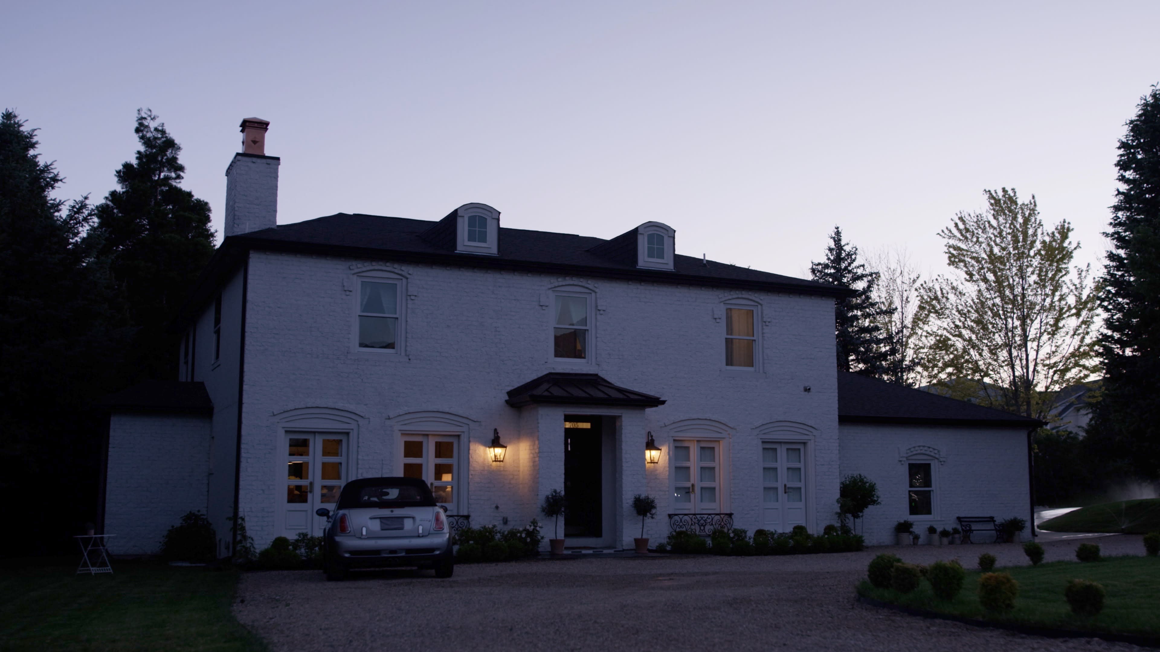 A white two-story house with a gravel driveway and a car parked outside is illuminated by soft exterior lights at dusk.