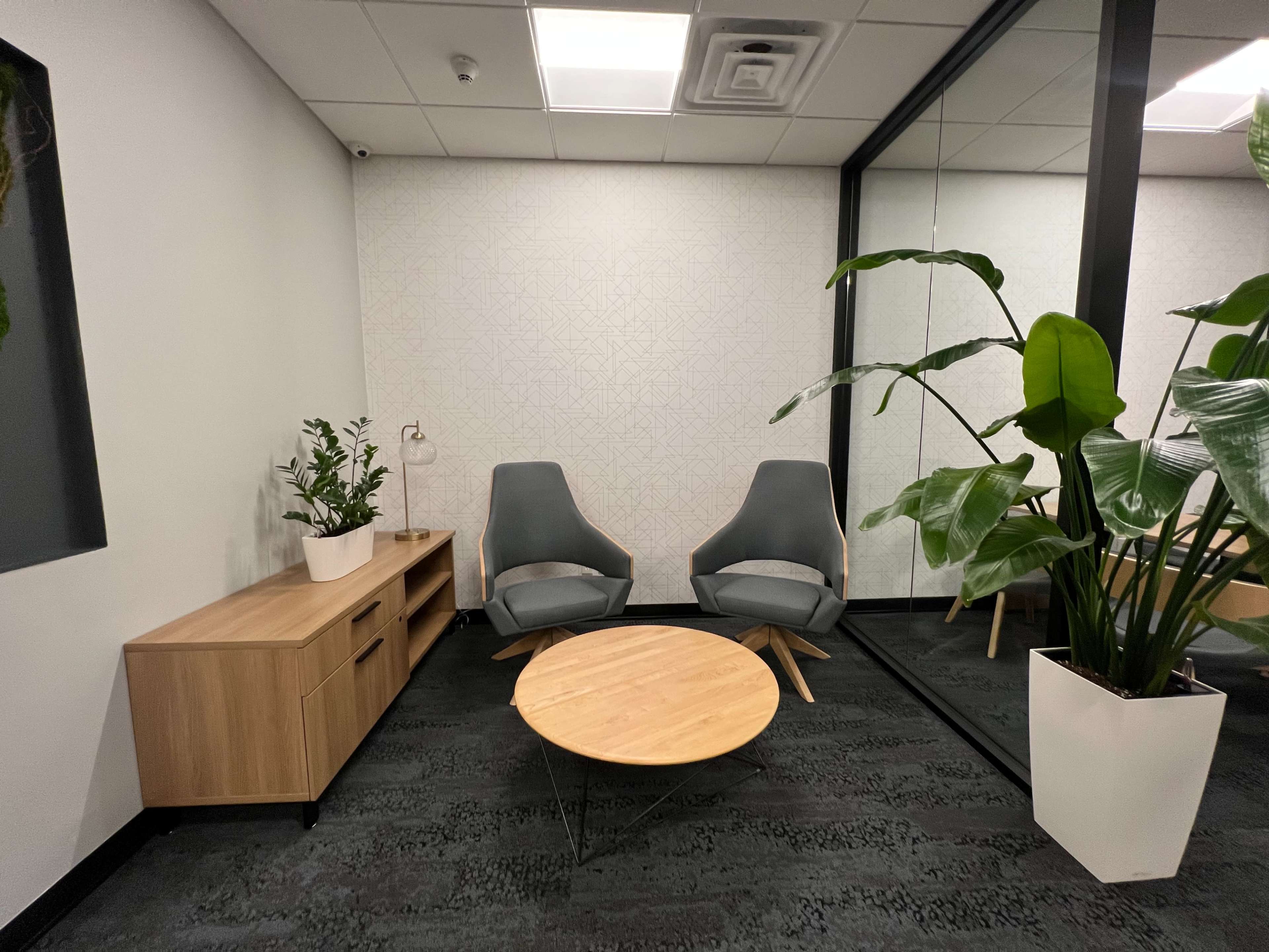A modern office lounge features two gray chairs, a wooden coffee table, and decorative plants against a light-colored wall and glass partition.