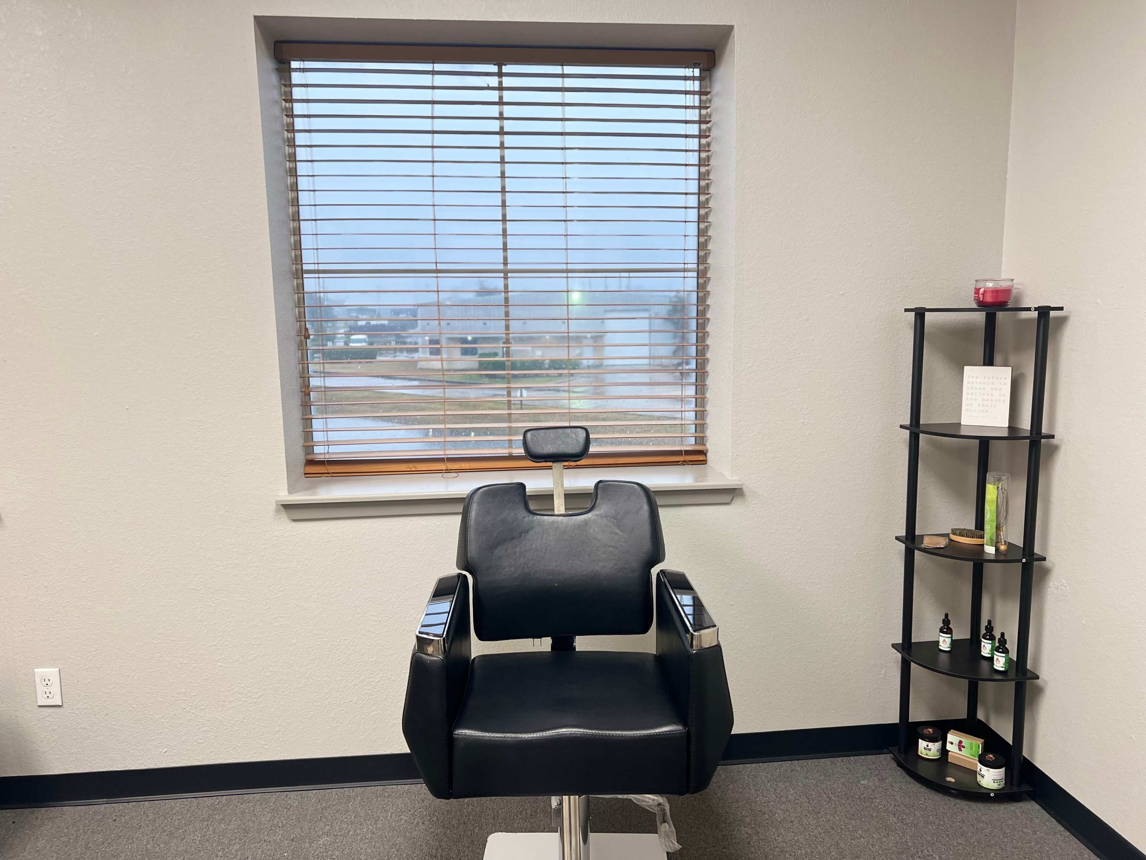 A black barber chair in a minimalist room with a window featuring blinds and a small shelf displaying various products.
