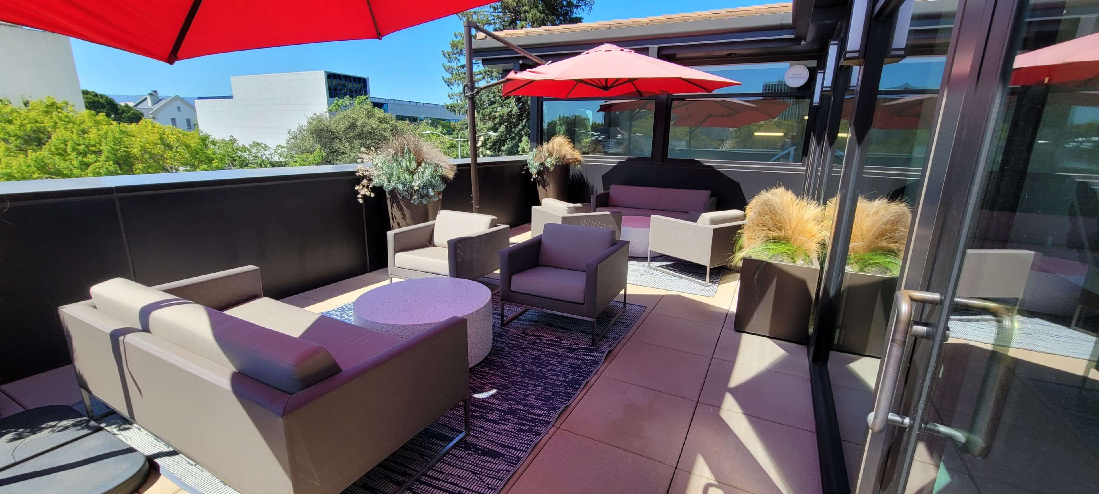 The image shows a rooftop terrace with several modern seating arrangements, a circular table, and red umbrellas providing shade.