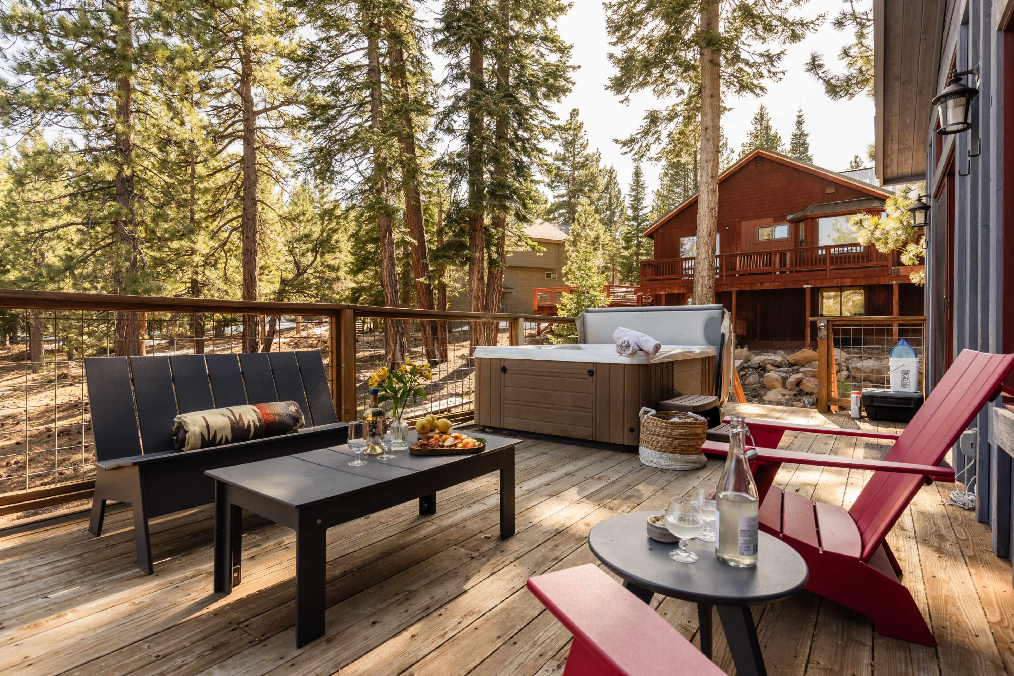 The image shows a wooden deck with outdoor furniture, including a dining table and chairs, adjacent to a hot tub, surrounded by tall trees and a cabin in the background.