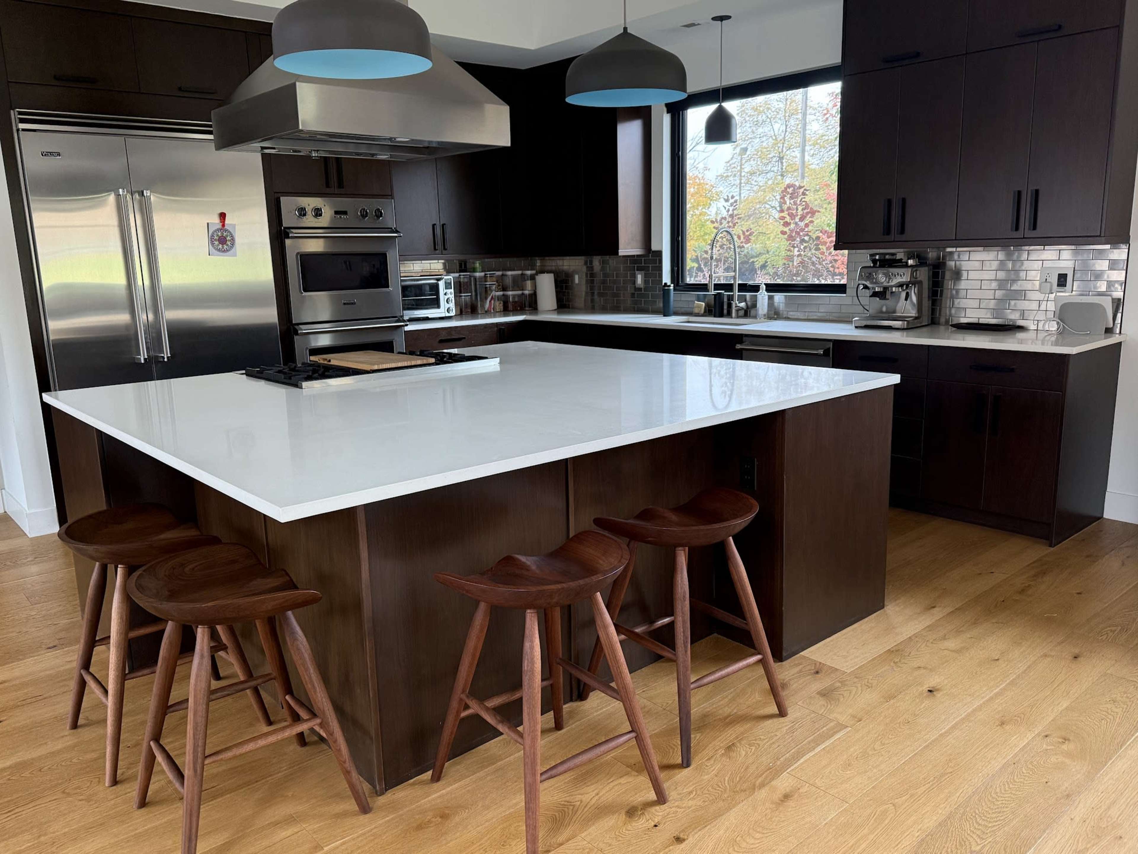 A modern kitchen features dark wood cabinetry, stainless steel appliances, a large white countertop island, and three wooden stools.