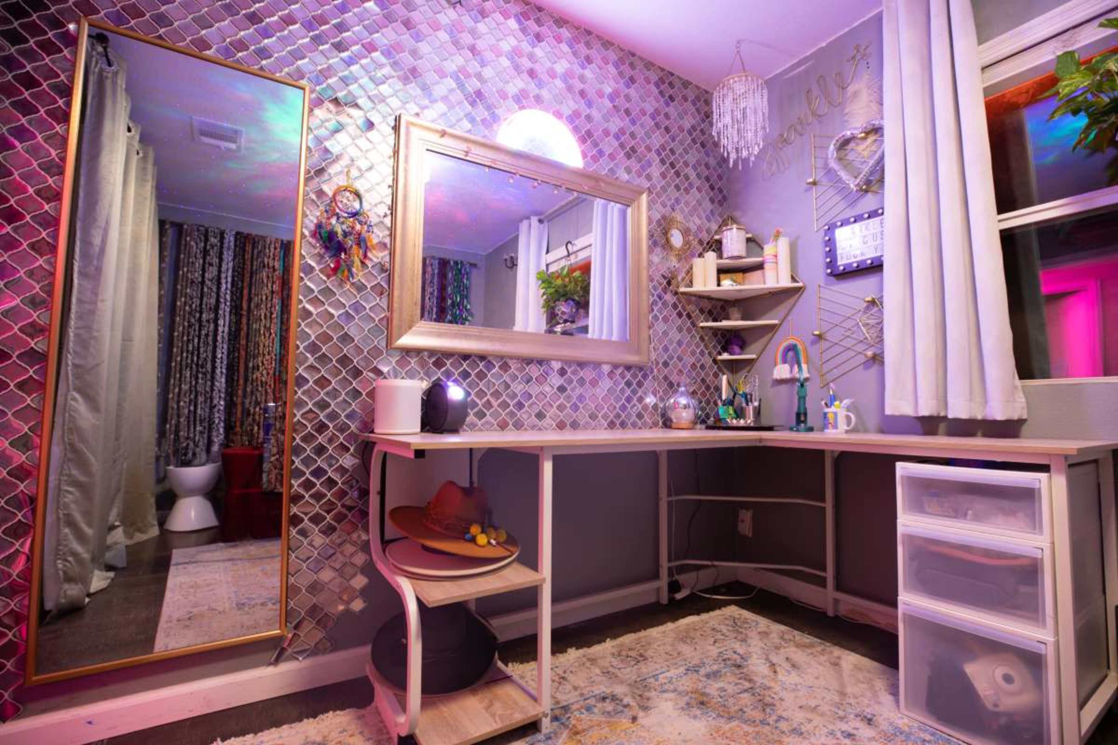 The image shows a modern vanity area with a large mirror, decorative wall tiles, a wooden desk, and colorful lighting.