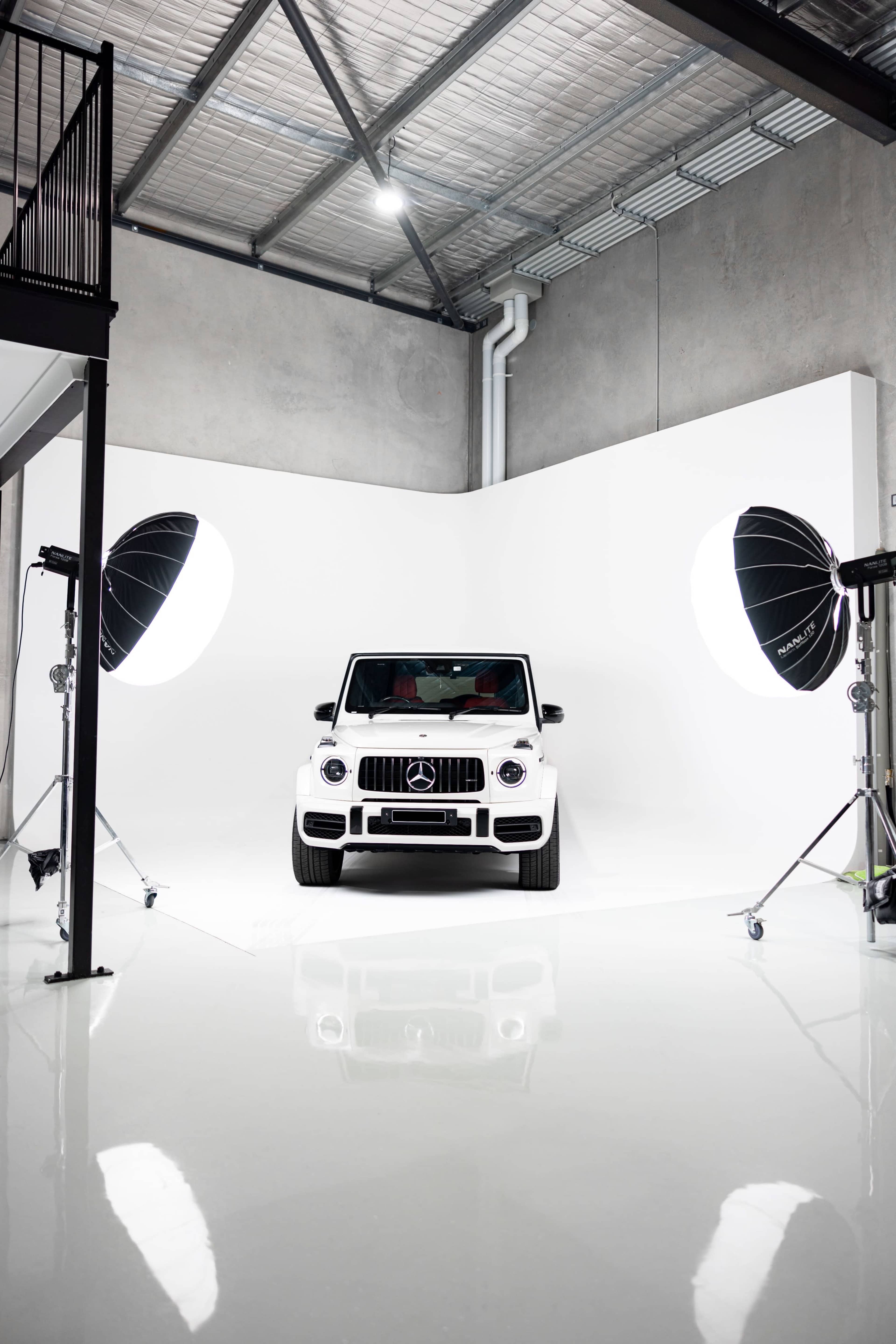 A white Mercedes-Benz G-Class is positioned in a well-lit studio with large softbox lights on either side and a seamless white backdrop.