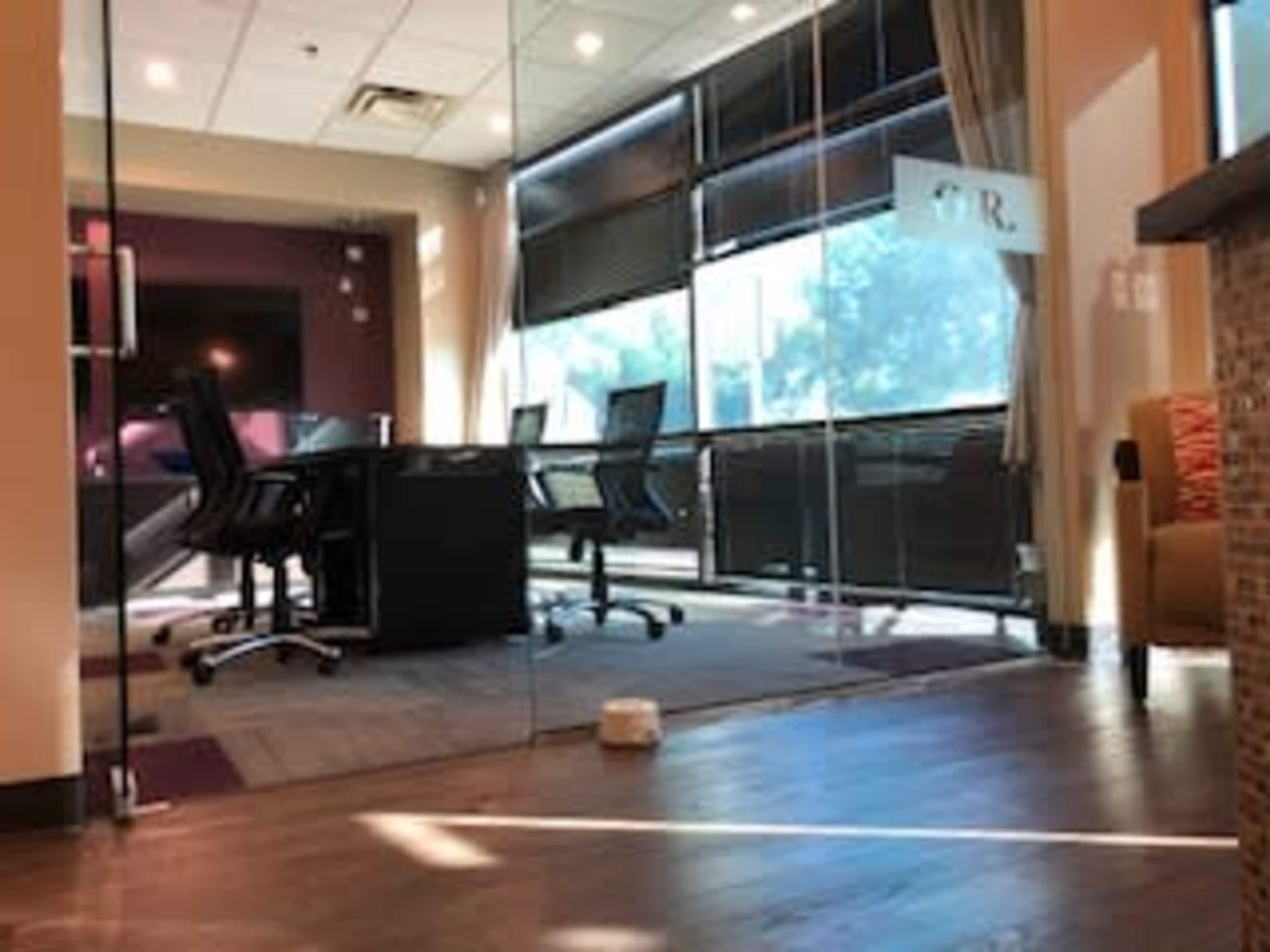 A modern office space with a glass-walled conference room containing a black table and rolling chairs, illuminated by natural light.